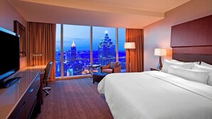 A hotel room with a large bed, a desk with a TV, a chair, and a view of a cityscape through large windows.