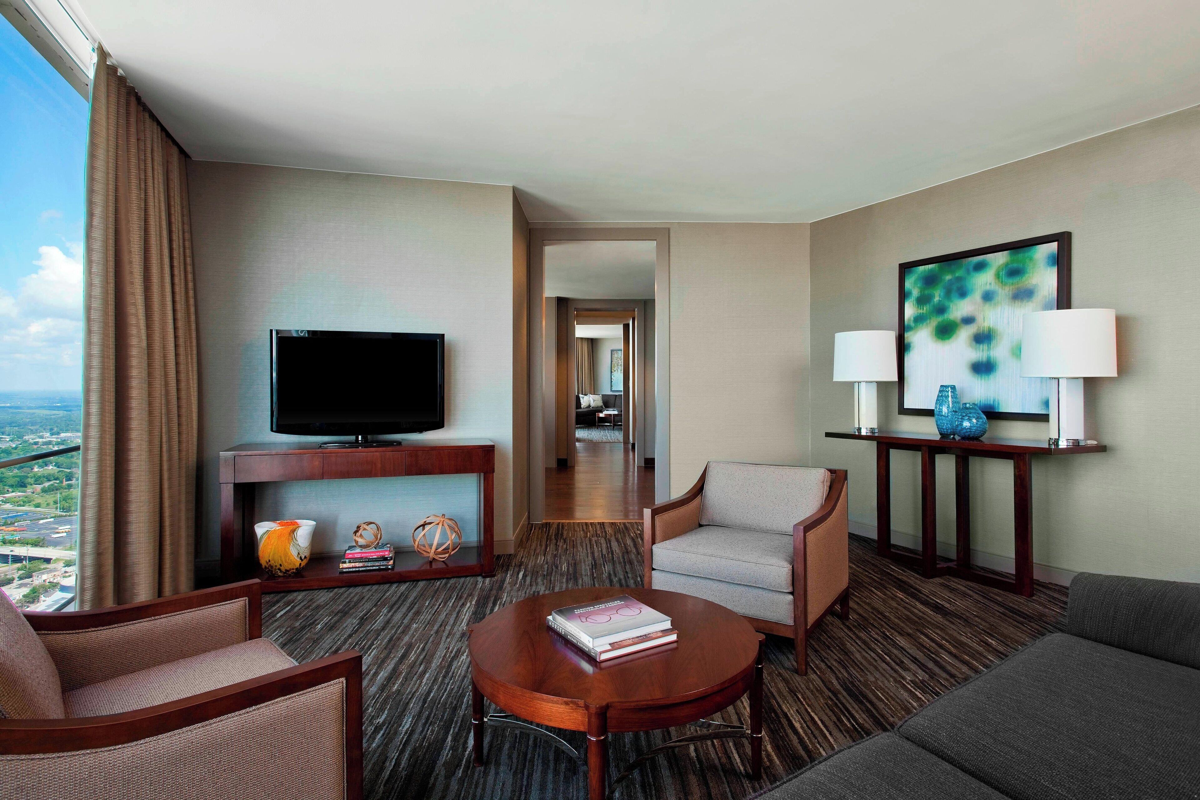 A hotel room with a television, a sofa, a coffee table, and a view of the city.