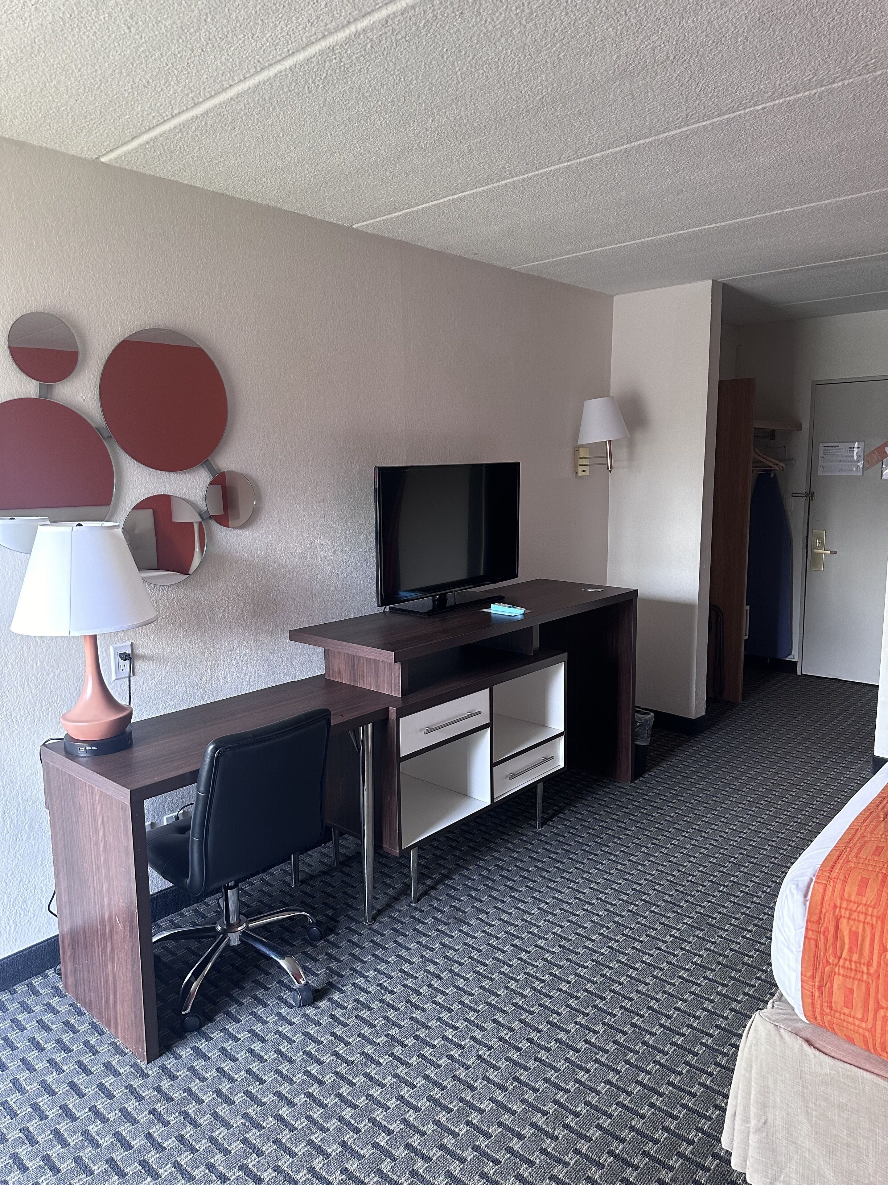Quarto de hotel com escrivaninha, cadeira, televisão, luminária e uma cama.