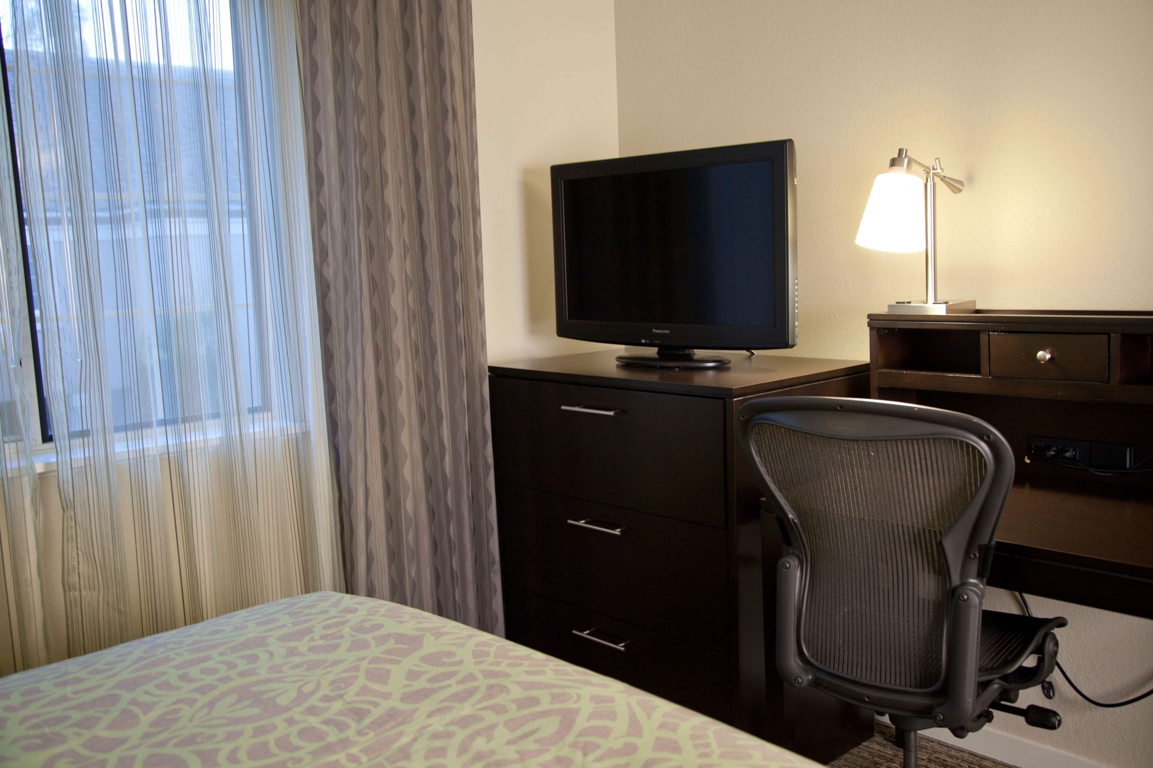 A hotel room with a bed, a desk with a television, a chair, and a lamp.