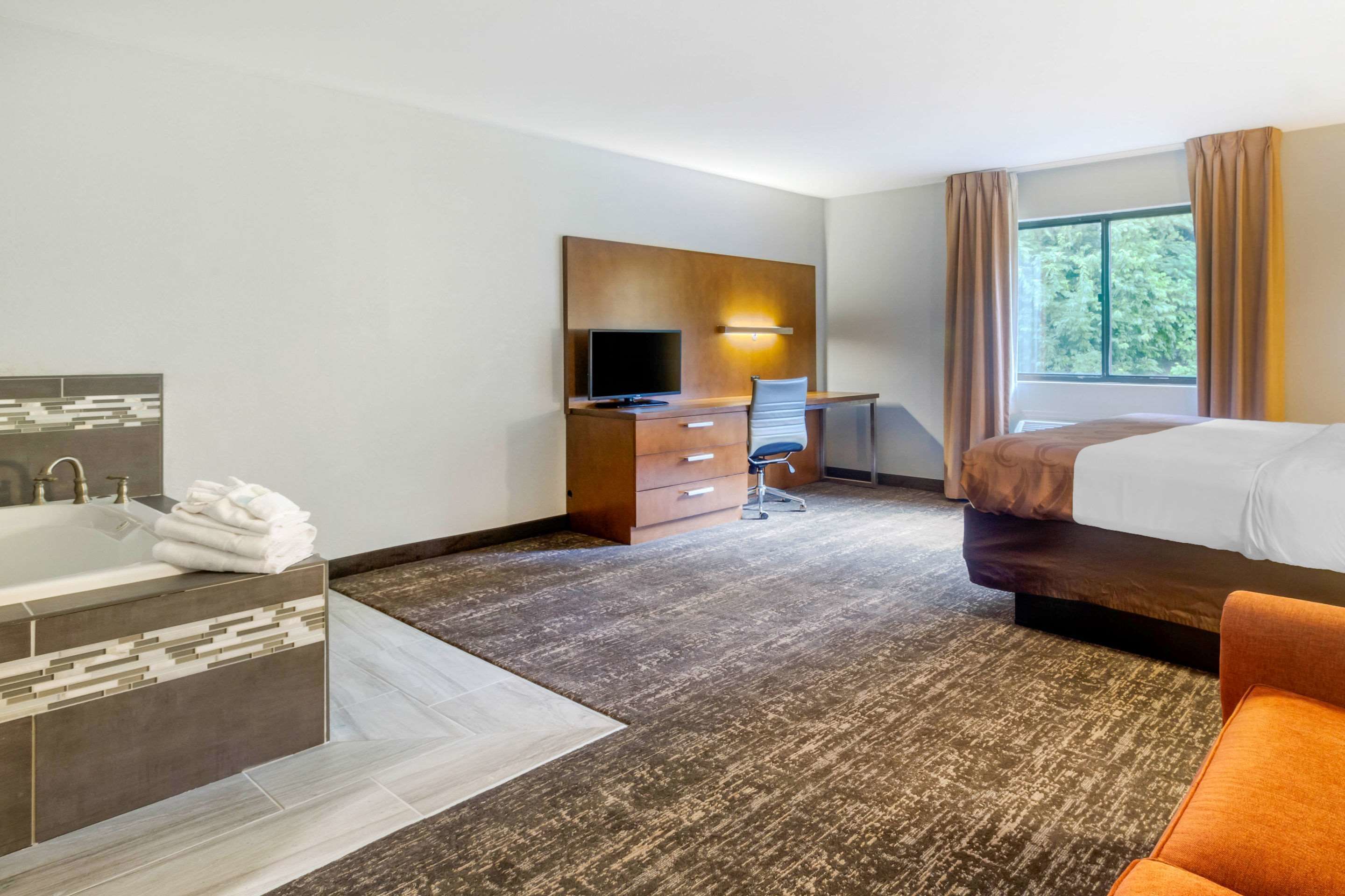 A hotel room with a large bed, a desk with a chair, a TV, a bathroom with a bathtub, and a window with curtains.
