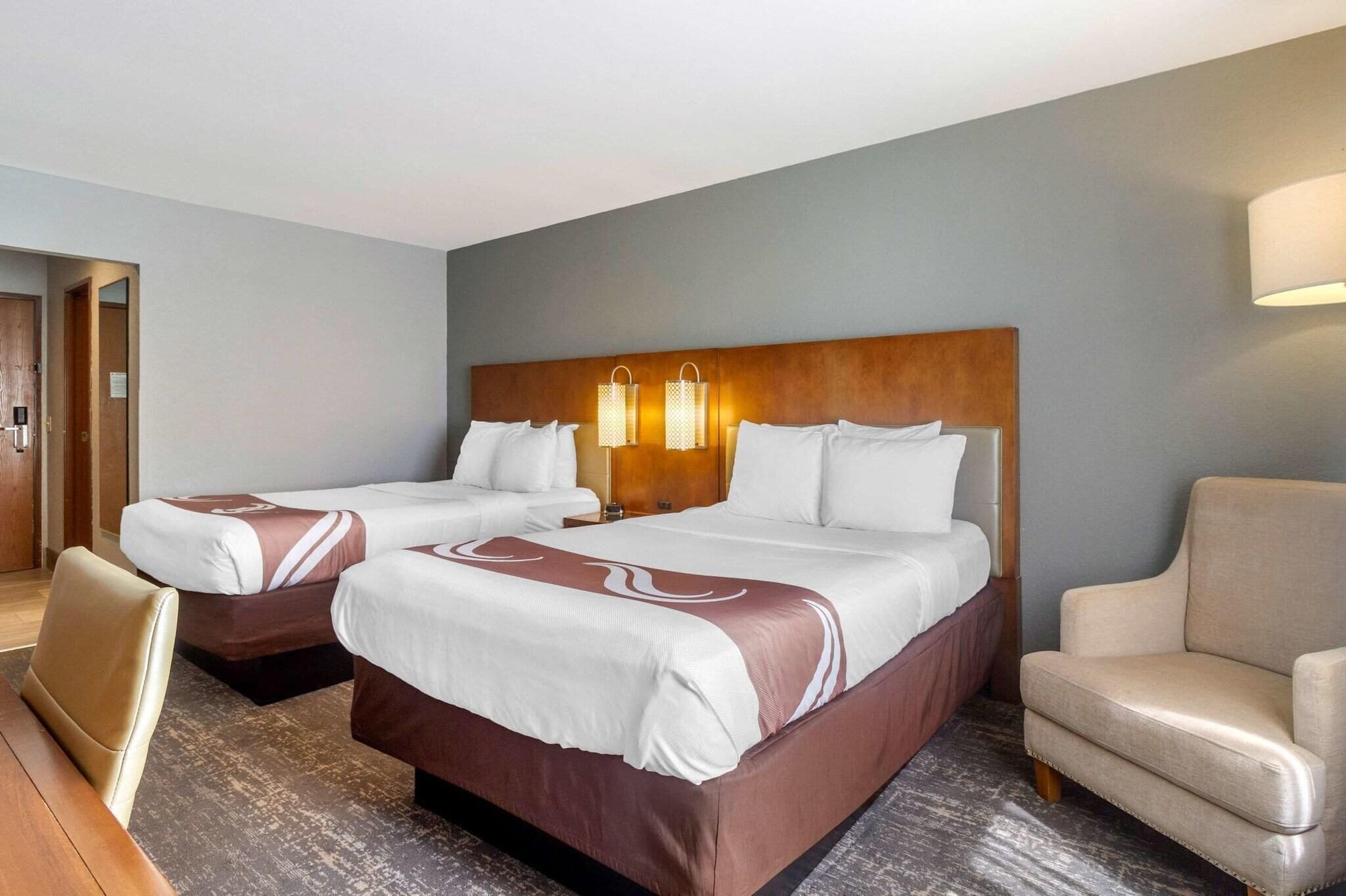 A hotel room with two beds, a wooden headboard, a desk, and a chair.