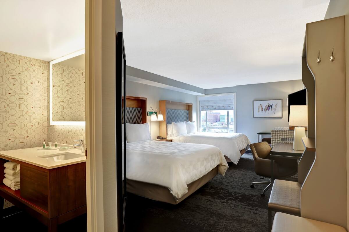 A hotel room with two beds, a desk, a chair, a desk lamp, a nightstand, a mirror, a sink, and a towel rack.