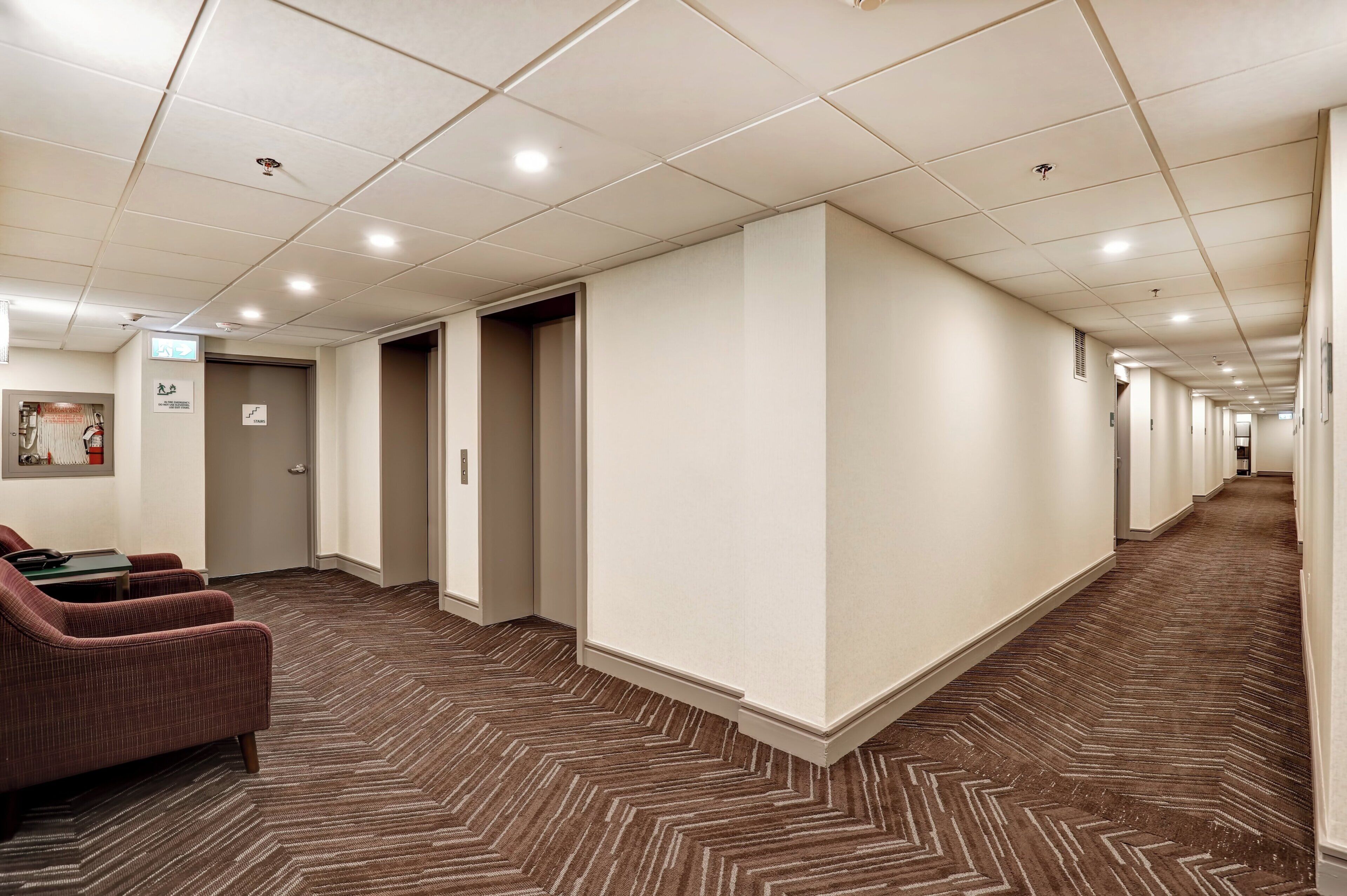 A hotel corridor with a herringbone-patterned carpet, a seating area with chairs, and a door marked 'Stairs'.