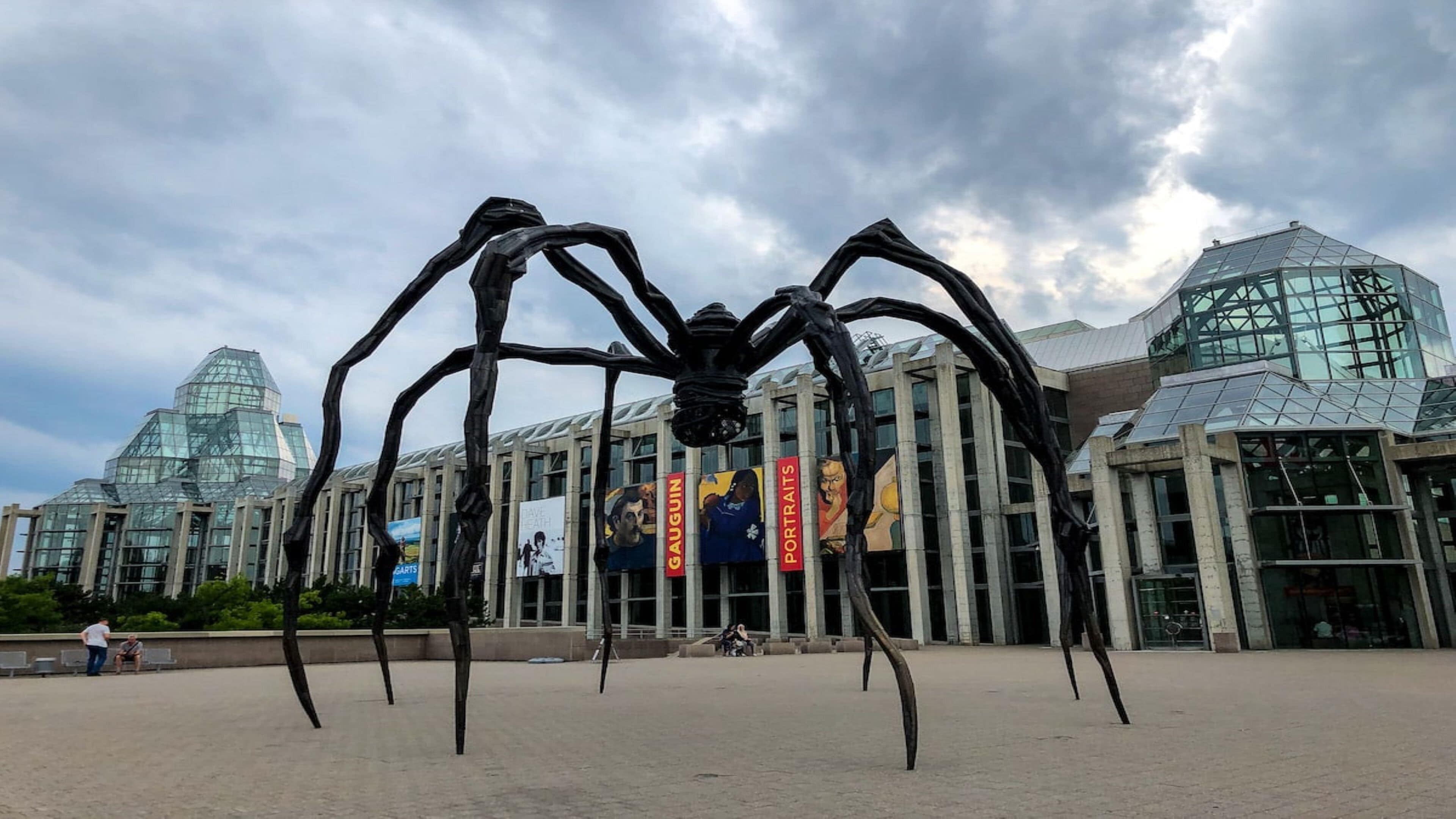 Une grande sculpture représentant une araignée en métal, placée devant un bâtiment moderne à l’architecture de verre et de béton.
