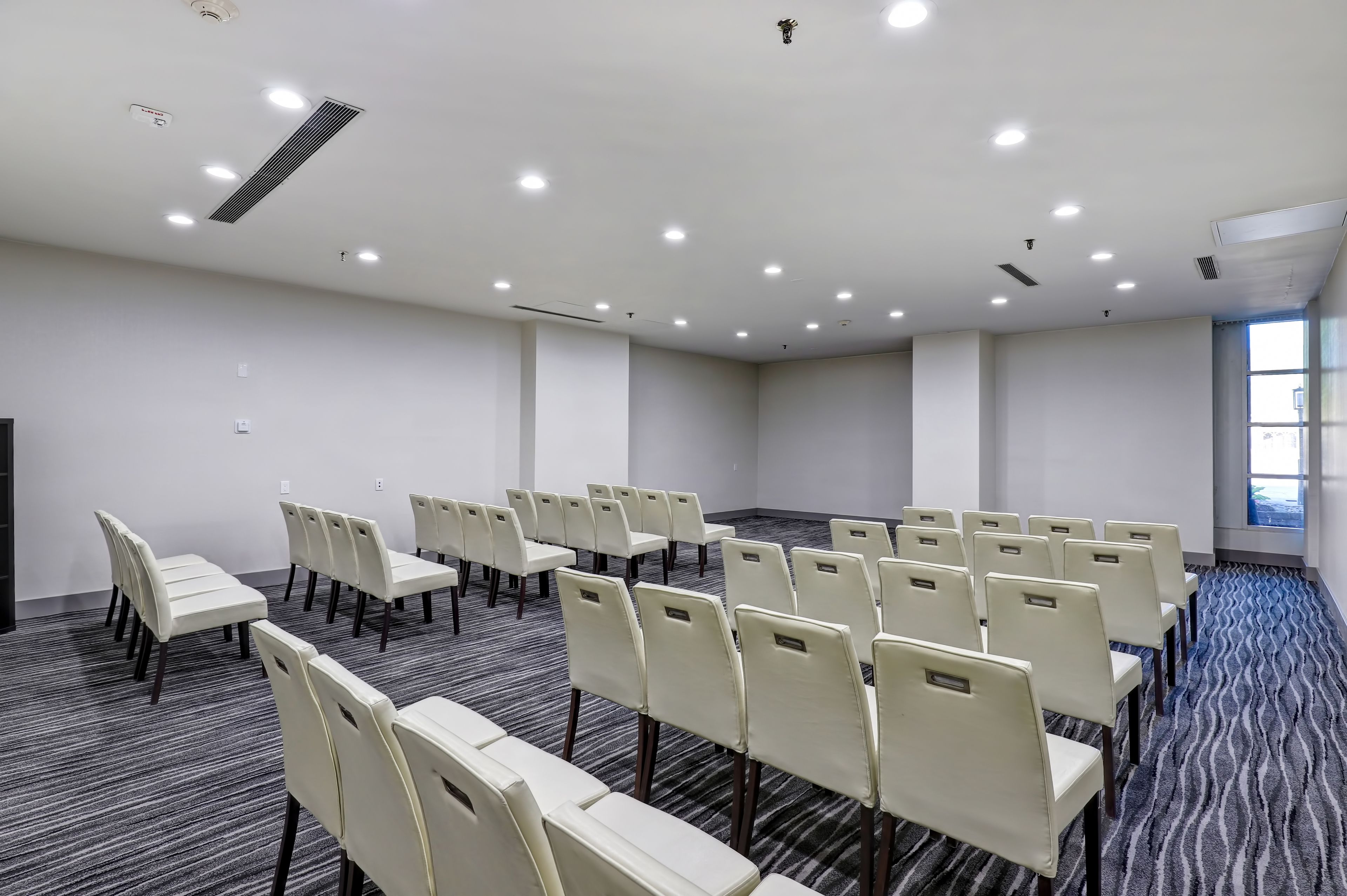 Un salón de conferencias con filas de sillas, piso alfombrado y un amplio ventanal.