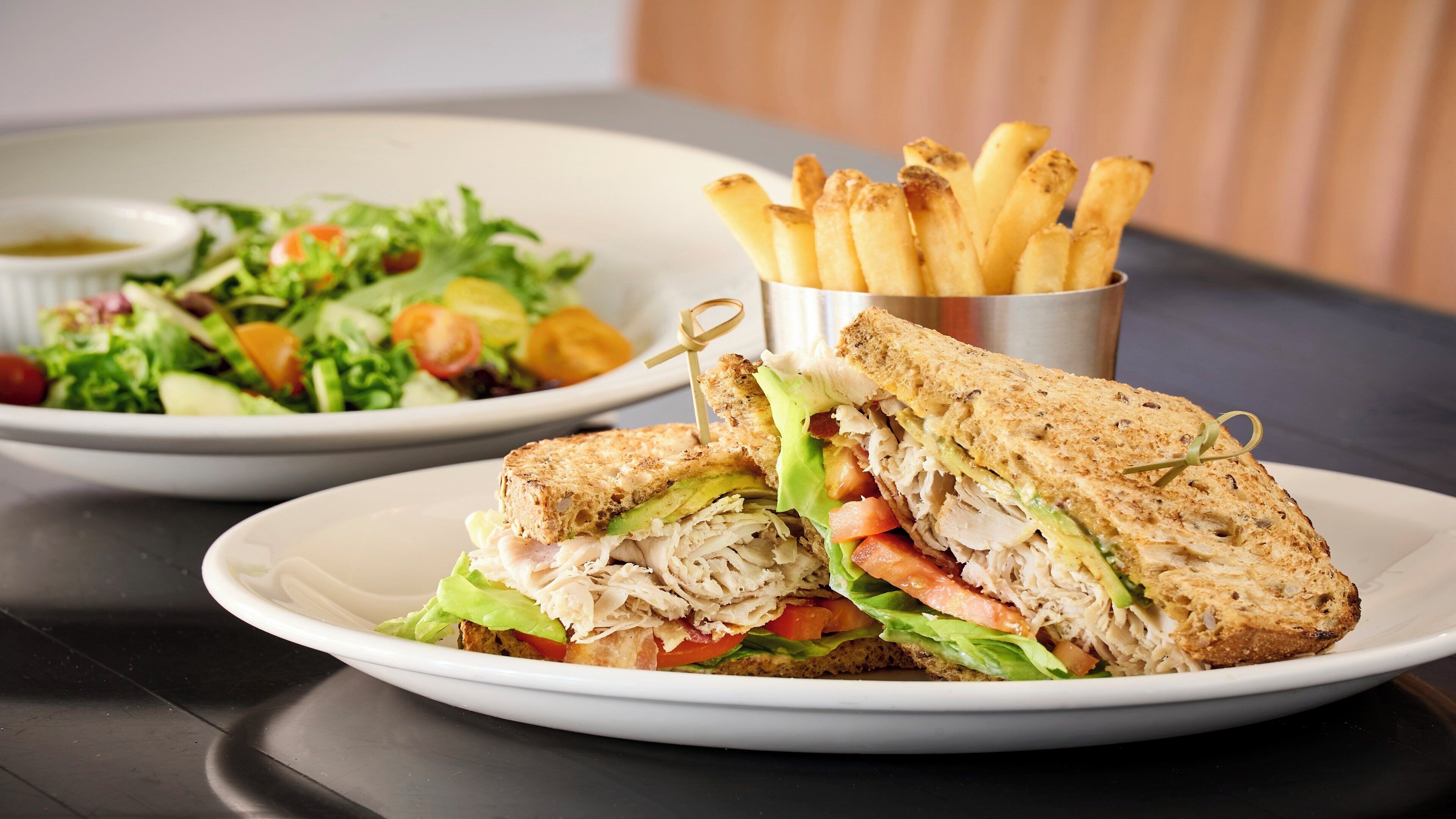 A sandwich cut in half on a plate, a bowl of salad, and a container of fries.