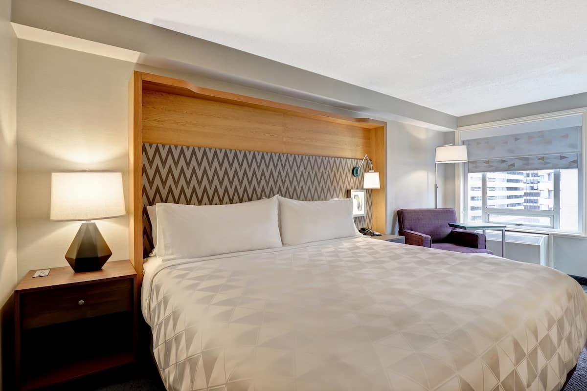 A hotel room with a large bed, wooden headboard, two bedside tables with lamps, a purple armchair, and a window with a view of buildings.