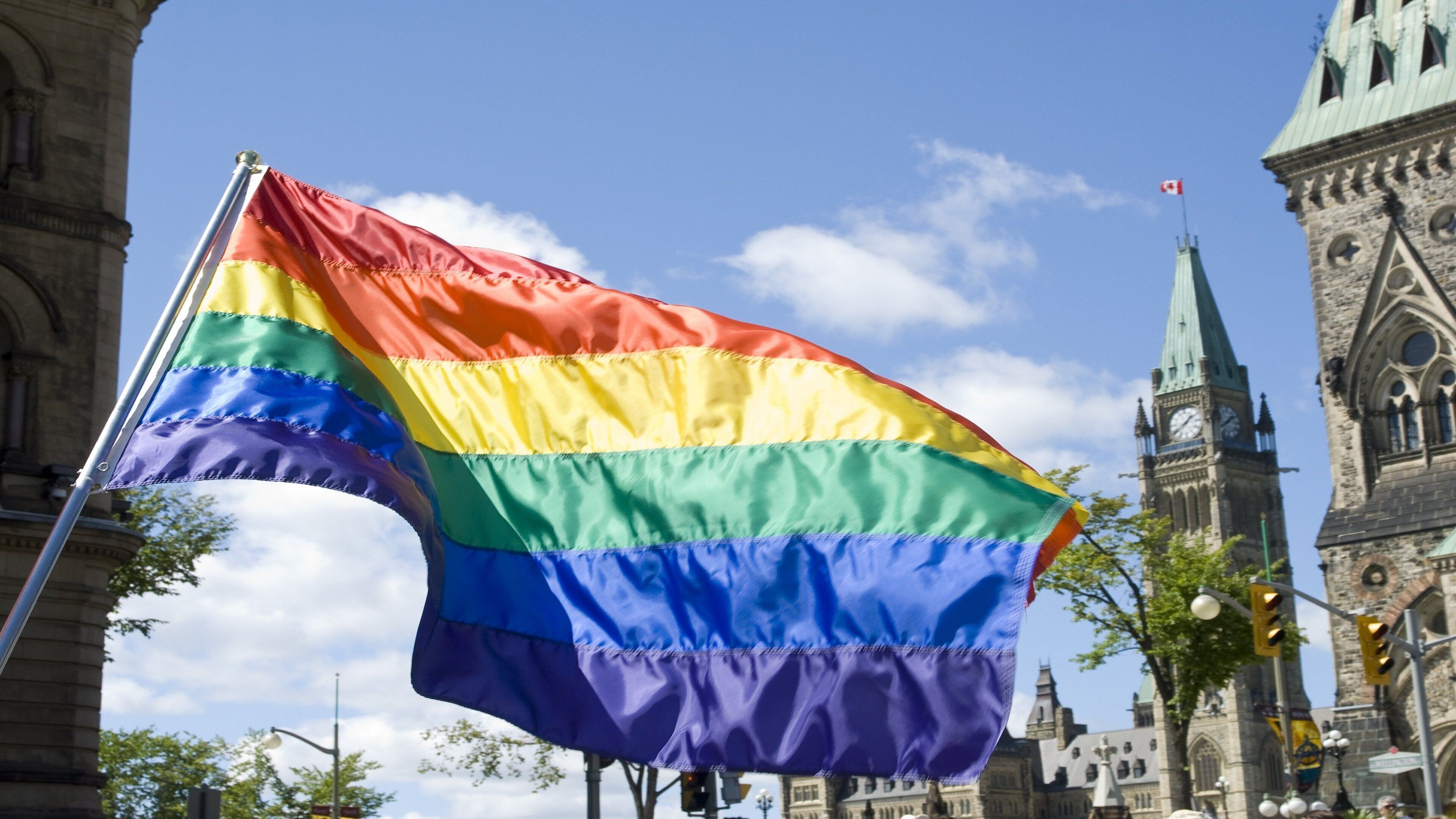 Un drapeau arc-en-ciel flotte devant un bâtiment historique doté d’une tour horloge.