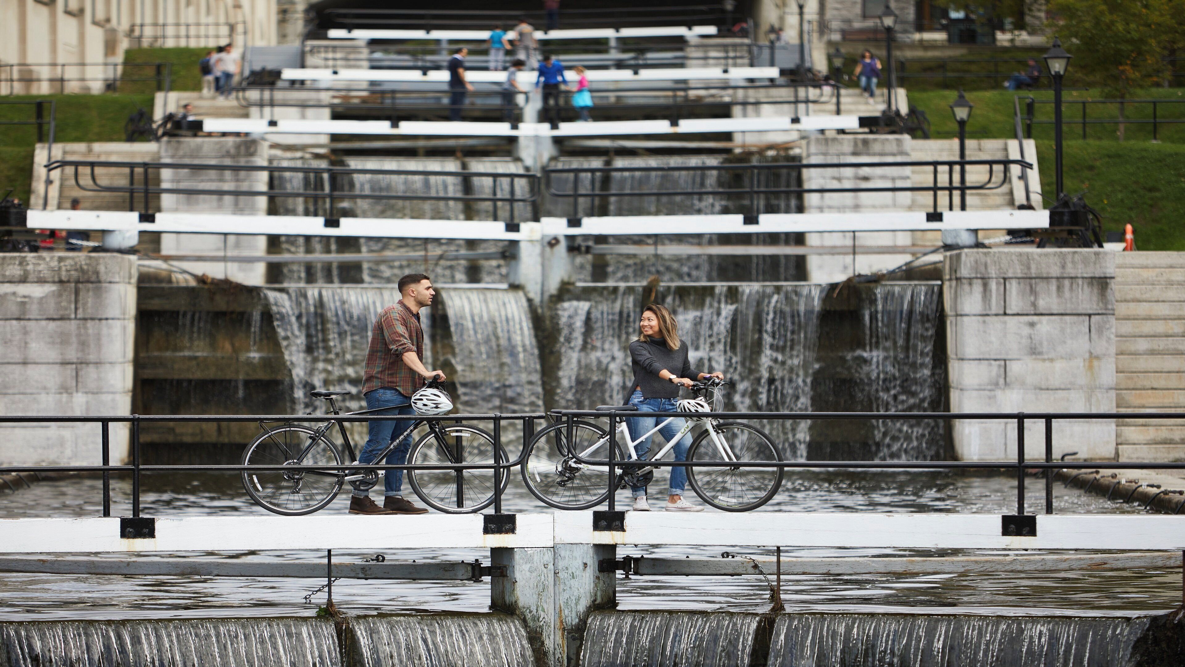 A man and a woman on bicycles near a waterfall with a stone structure in the background.