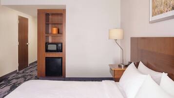 A hotel room with a bed, a wooden shelf unit with a microwave, a lamp, and a door.