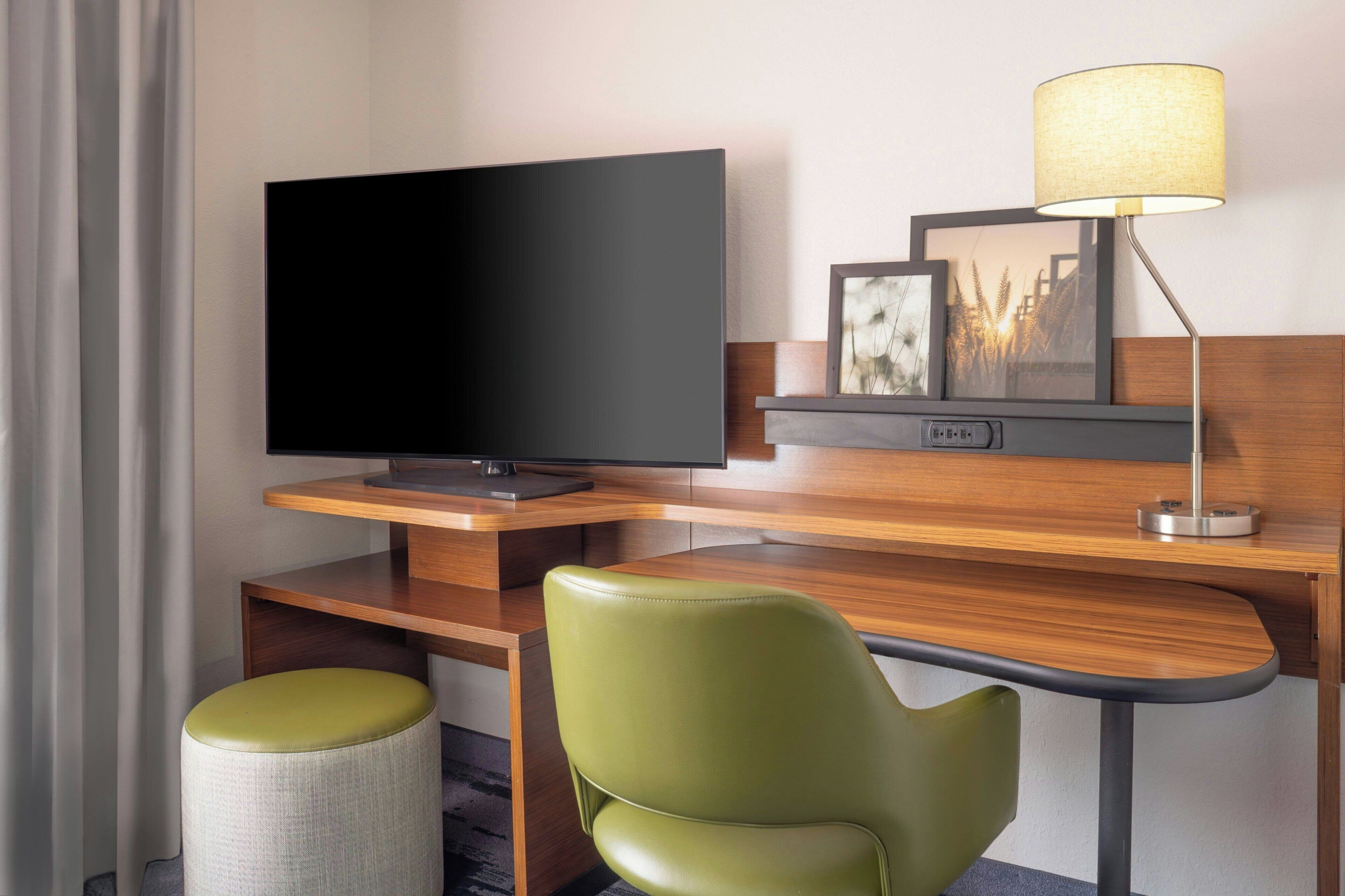 A hotel room with a flat-screen TV, a desk with a lamp, a green chair, and a small stool.