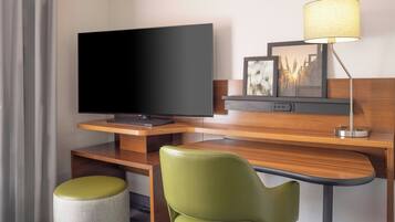 A hotel room with a flat-screen TV, a desk with a lamp, a green chair, and a small stool.