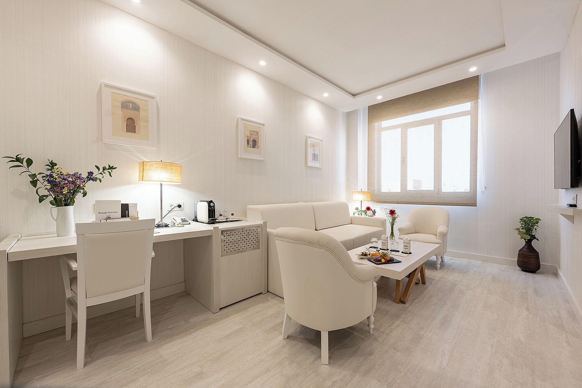 Habitación de hotel moderna con escritorio, sofá, sillón y mesa de comedor. Hay dos lámparas, un televisor y una ventana con cortinas.