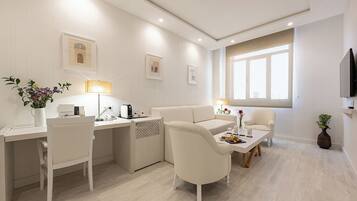 Habitación de hotel moderna con escritorio, sofá, sillón y mesa de comedor. Hay dos lámparas, un televisor y una ventana con cortinas.