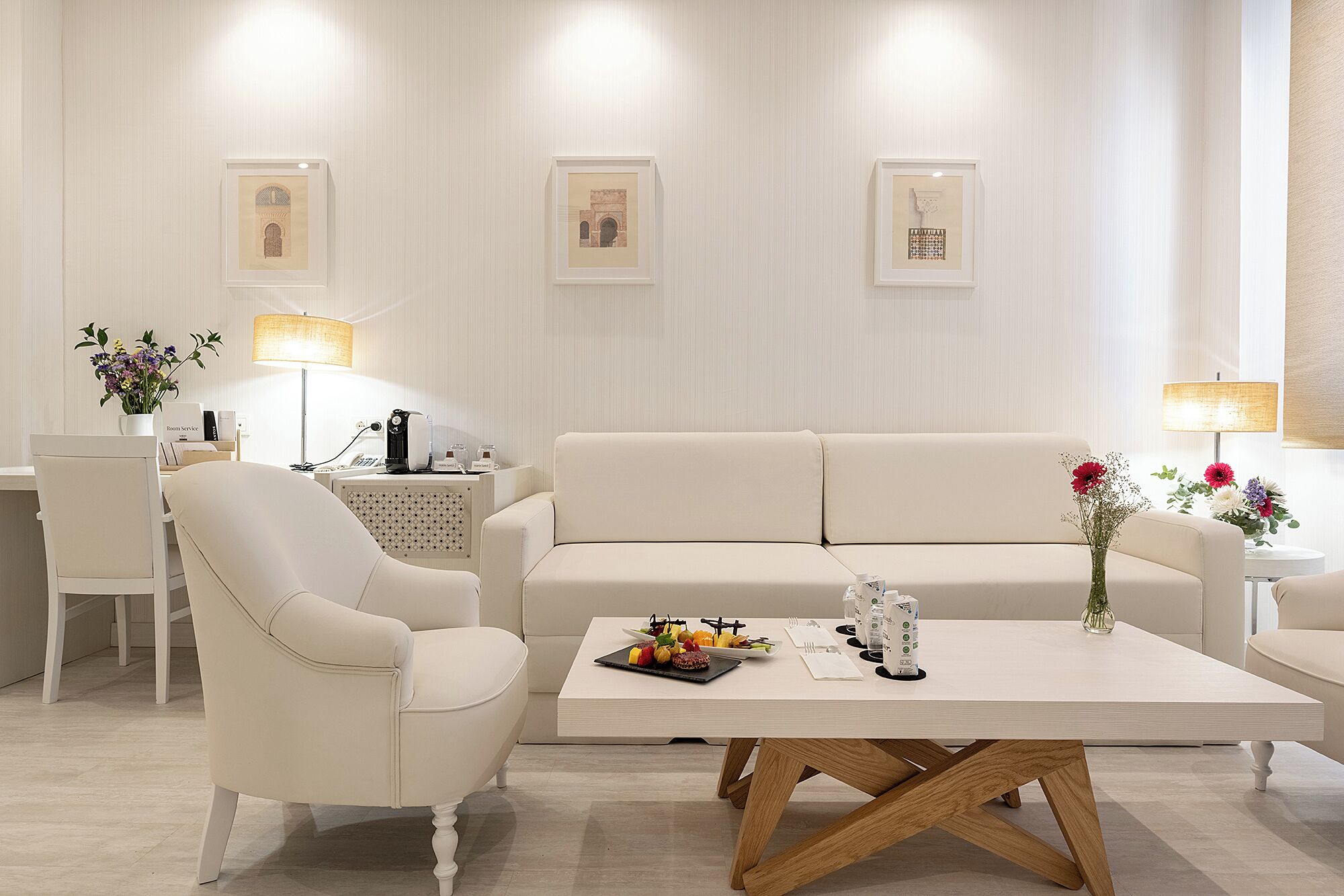 A modern hotel room with a white sofa, a dining table with a fruit platter, a coffee machine, and framed artwork on the wall.