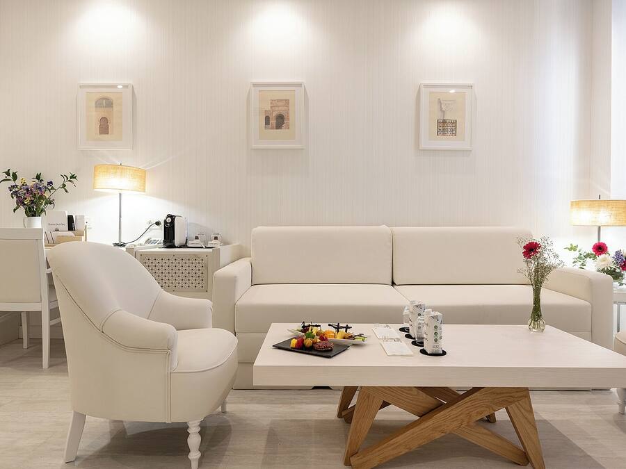 A modern hotel room with a white sofa, a dining table with a fruit platter, a coffee machine, and framed artwork on the wall.