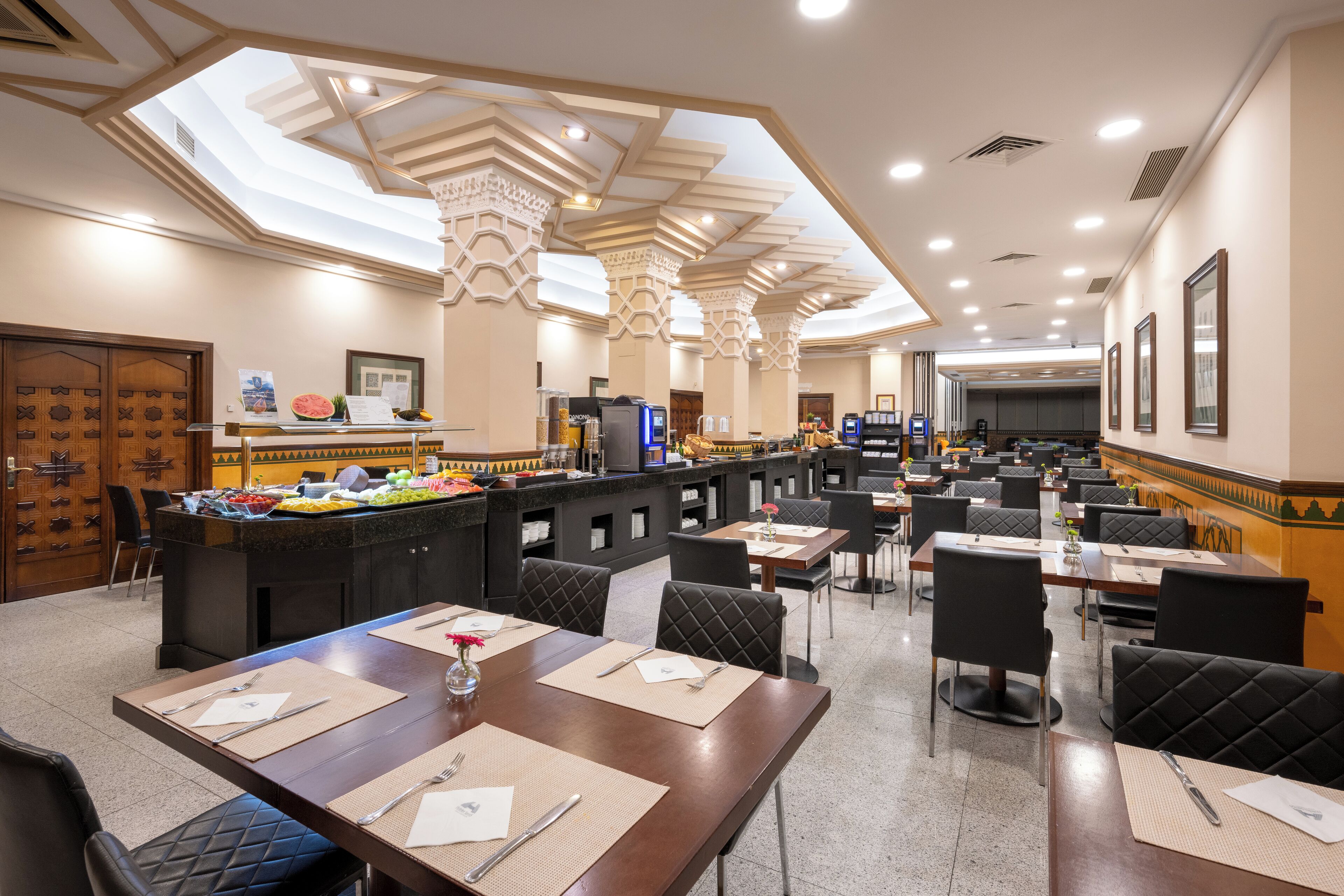 Un ristorante spazioso con numerosi tavoli e sedie, un'area bar centrale e un soffitto alto con finestre a lucernario.