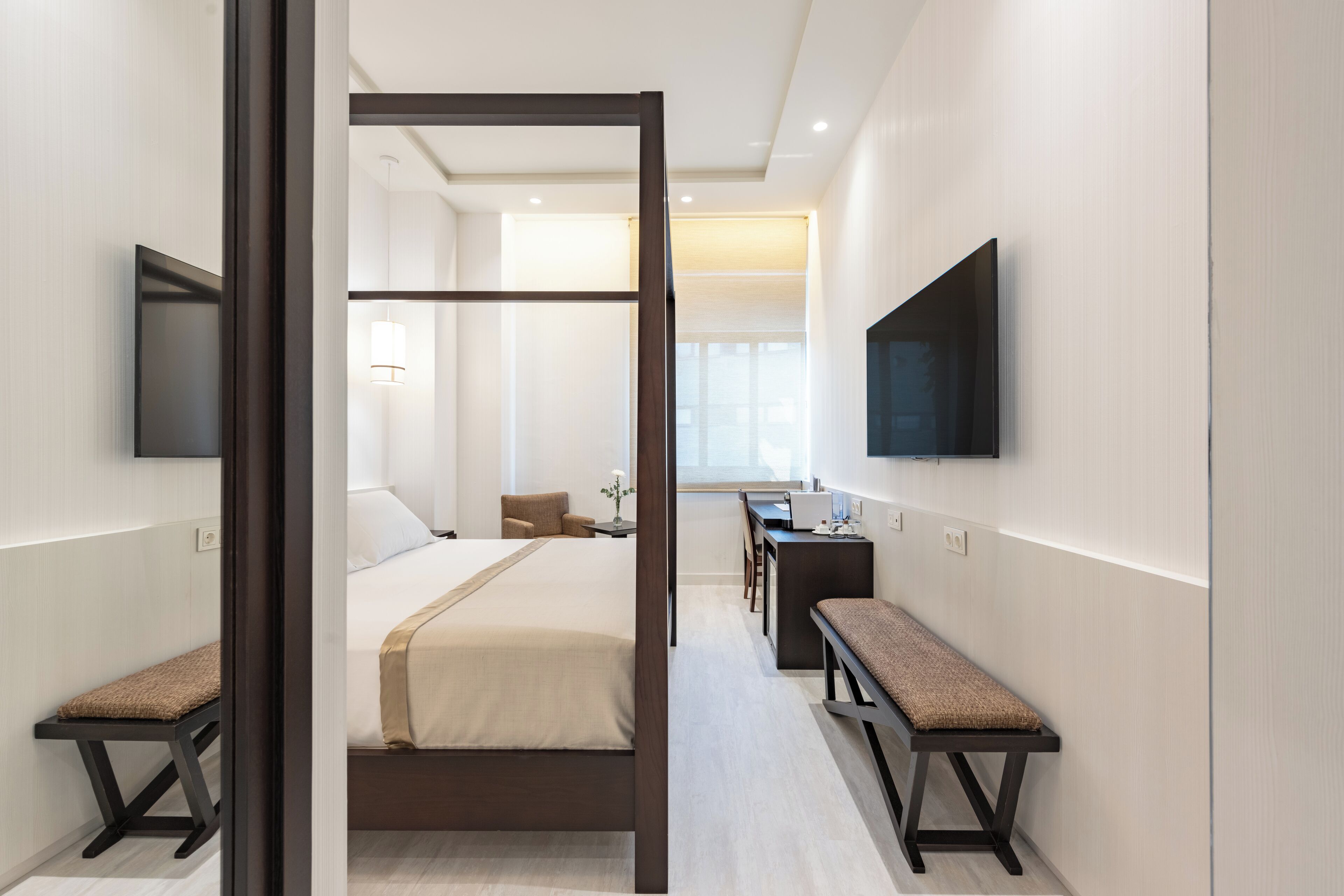 A modern hotel room with a large bed, a desk, a bench, and a TV mounted on the wall.