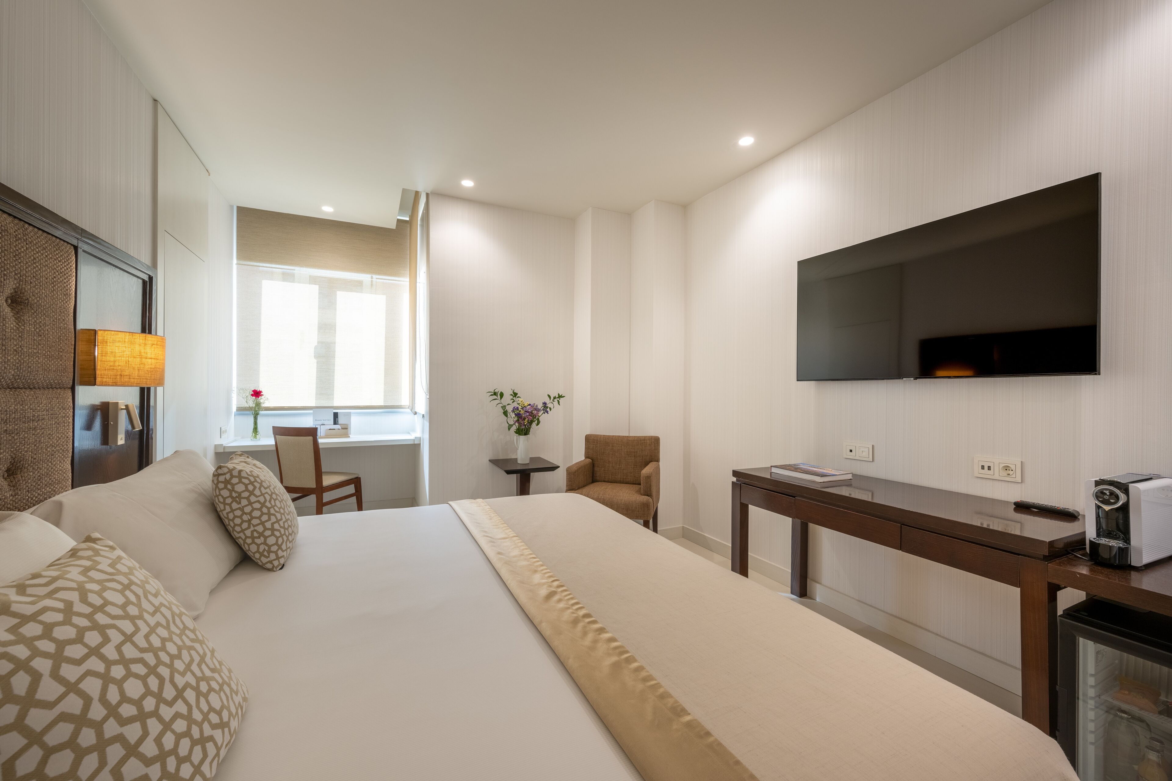 A hotel room with a large bed, a desk, a chair, a TV mounted on the wall, and a coffee machine.