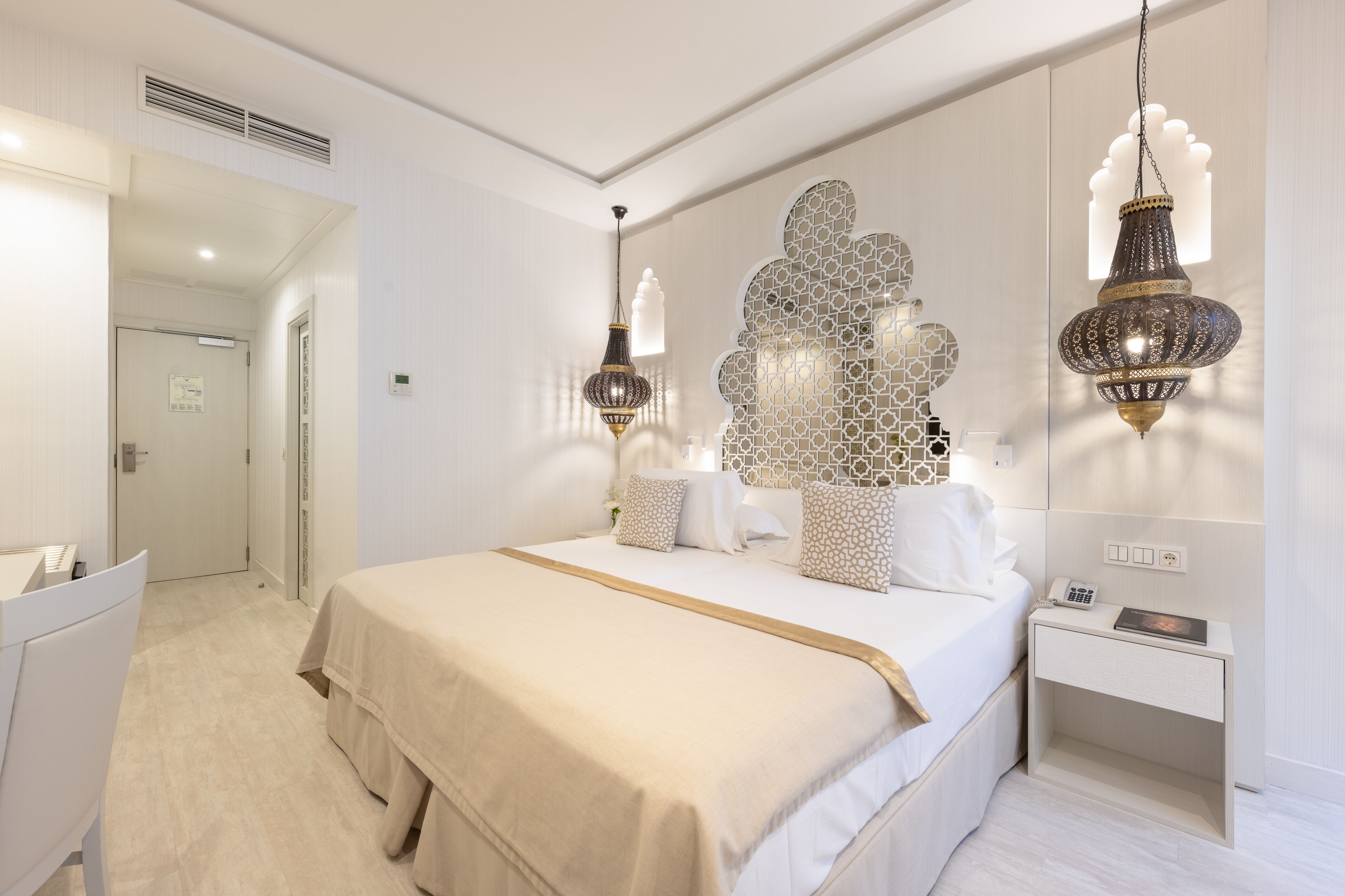 A hotel room with a large bed, bedside tables, a desk, and decorative wall sconces.