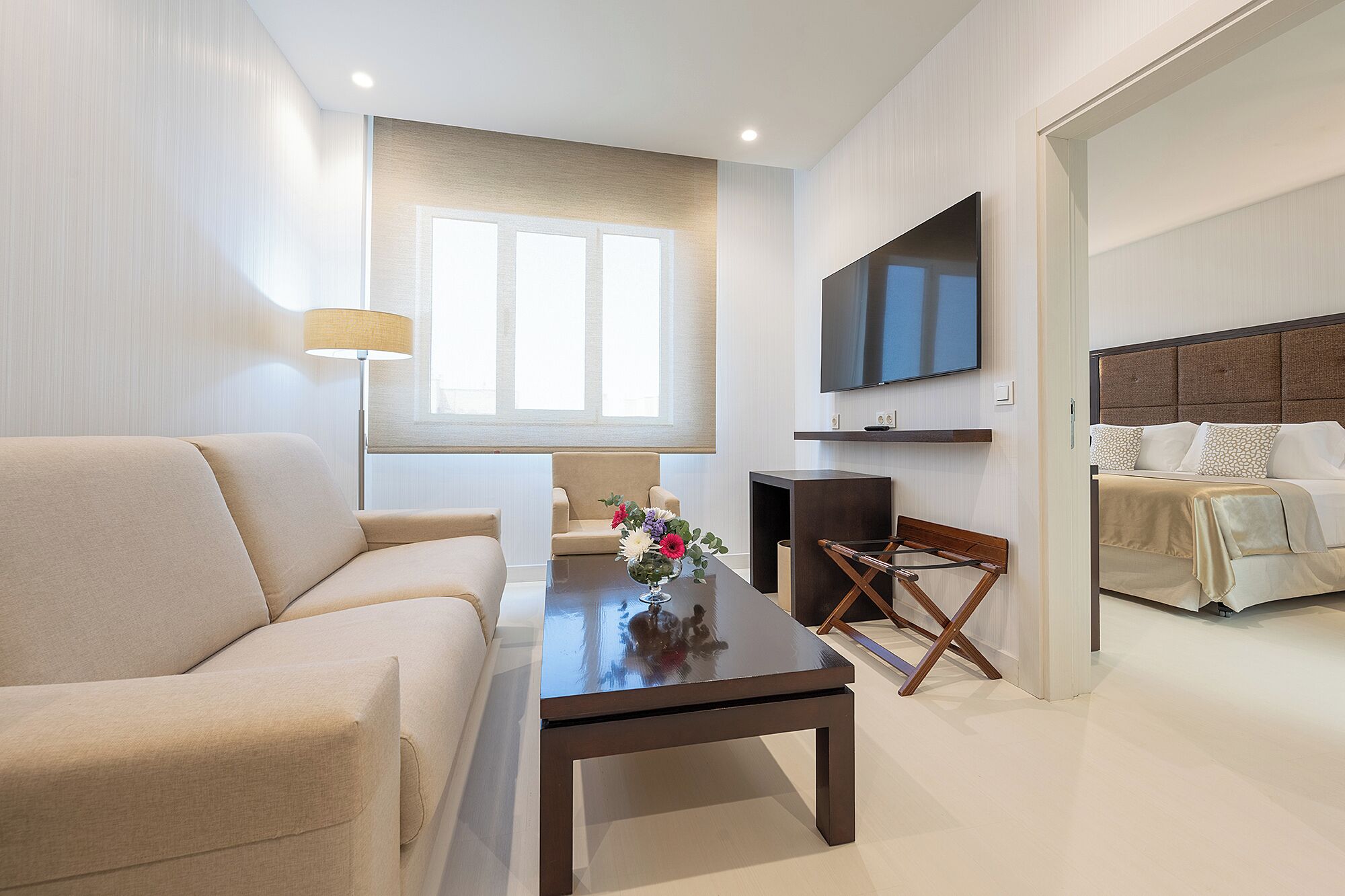 A modern living room with a sofa, a coffee table with flowers, a TV, and a bed in the background.