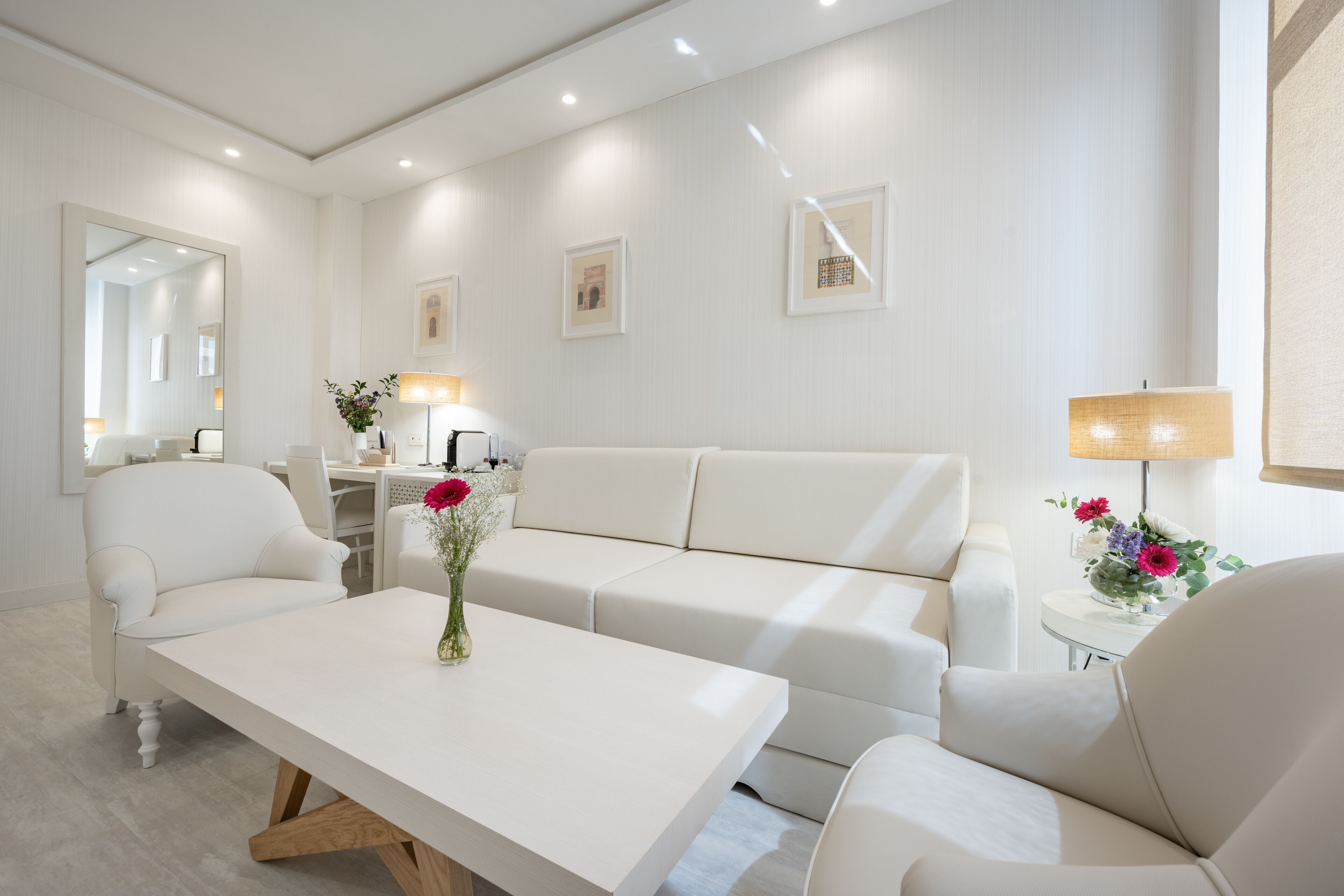 Un salón moderno y bien iluminado con un sofá blanco, una mesa de centro con un jarrón de flores y un comedor con una lámpara.