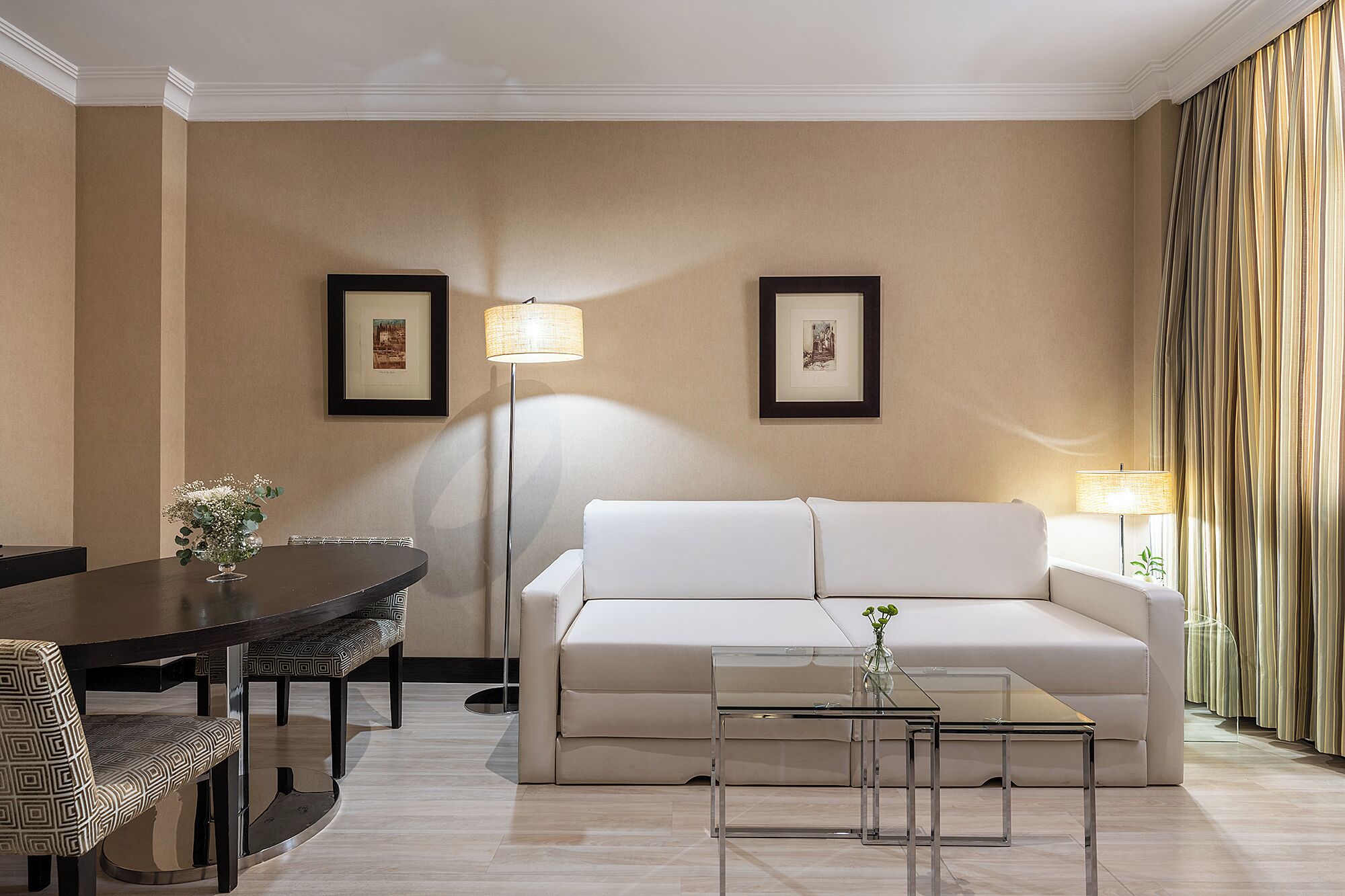 A modern living room with a sofa, coffee table, dining table, and chairs. There are two framed pictures on the wall and two floor lamps.