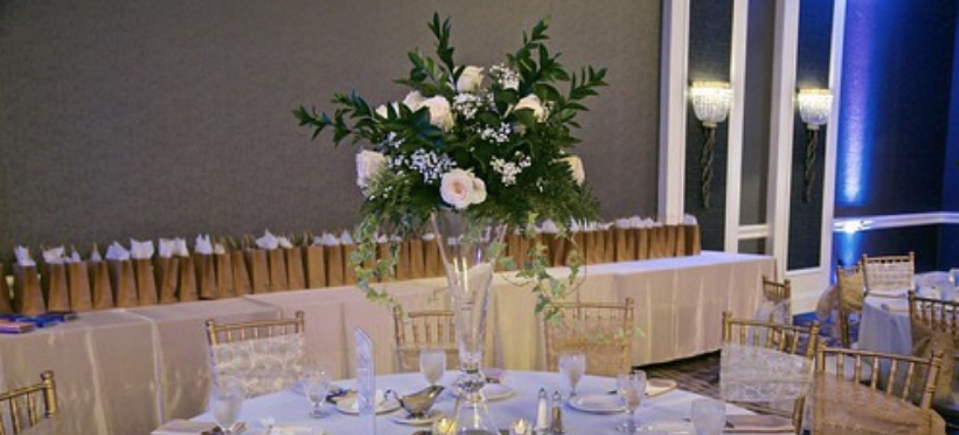 Una mesa de banquete con un centro de mesa floral, manteles blancos y sillas doradas.