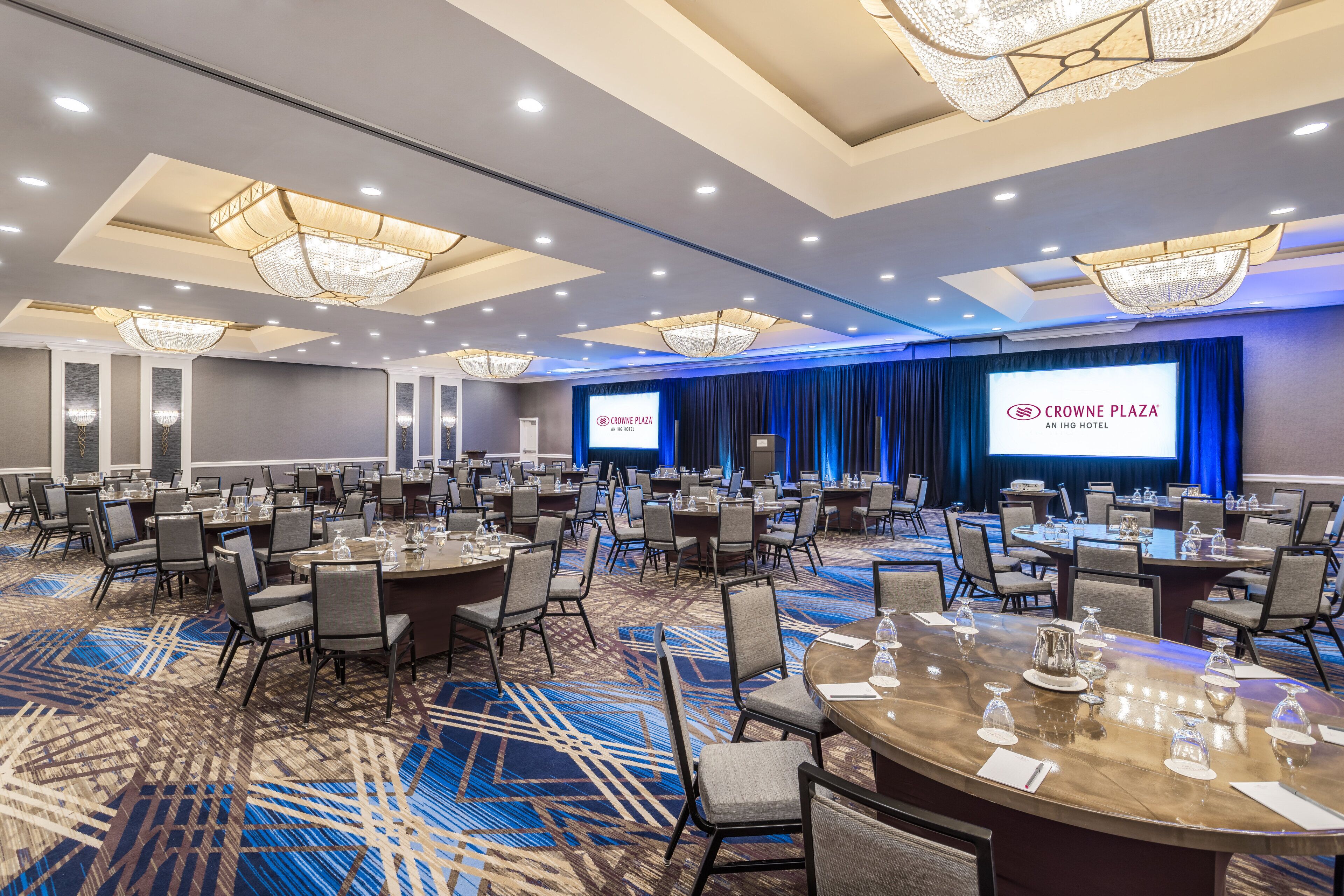 A conference room with multiple tables and chairs, a large screen displaying the hotel name, and a blue carpet.