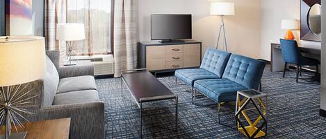 A hotel room with a sofa, a small table, a television, a desk with a chair, and a lamp.