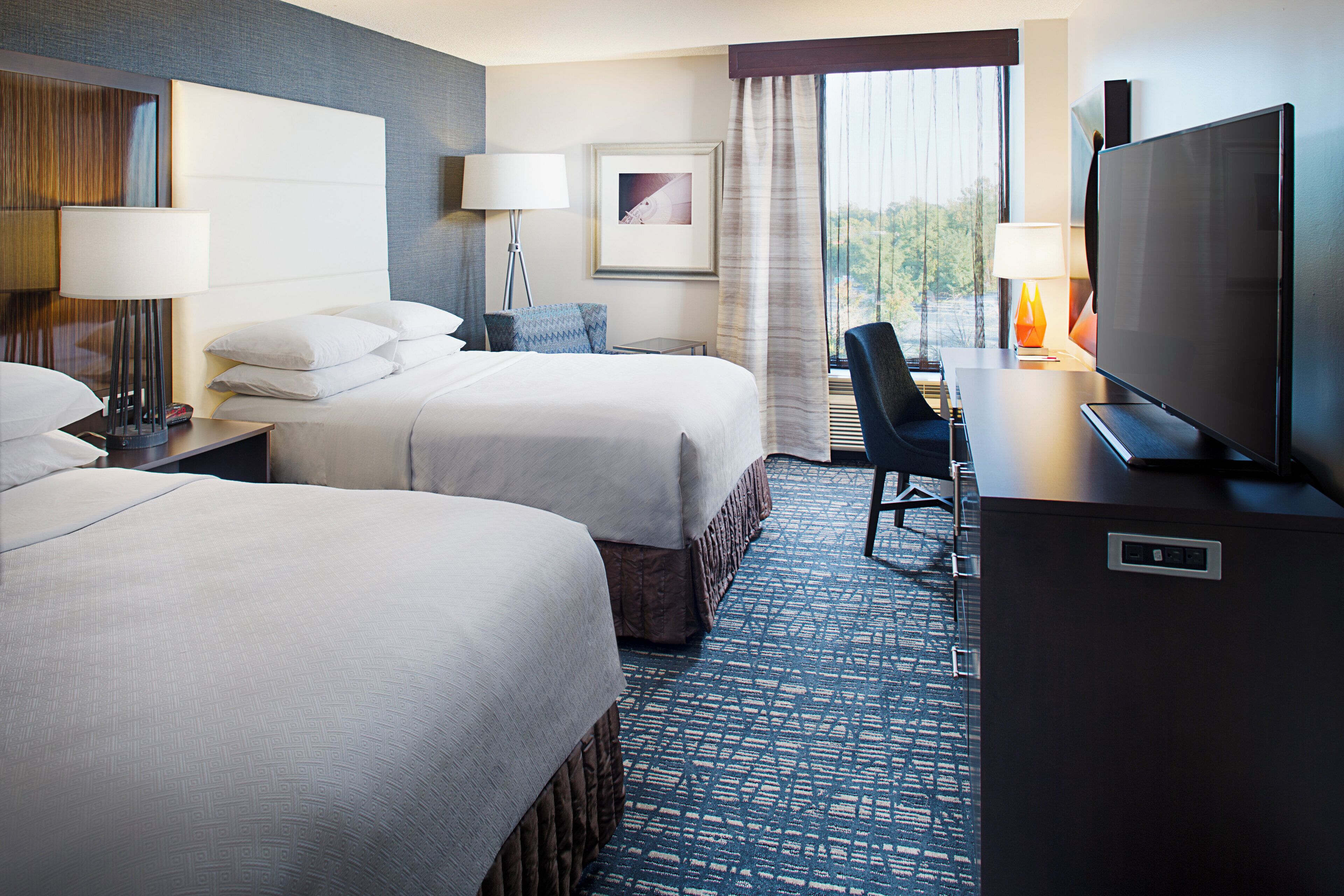 A hotel room with two beds, a desk with a television, a lamp, and a framed picture on the wall.