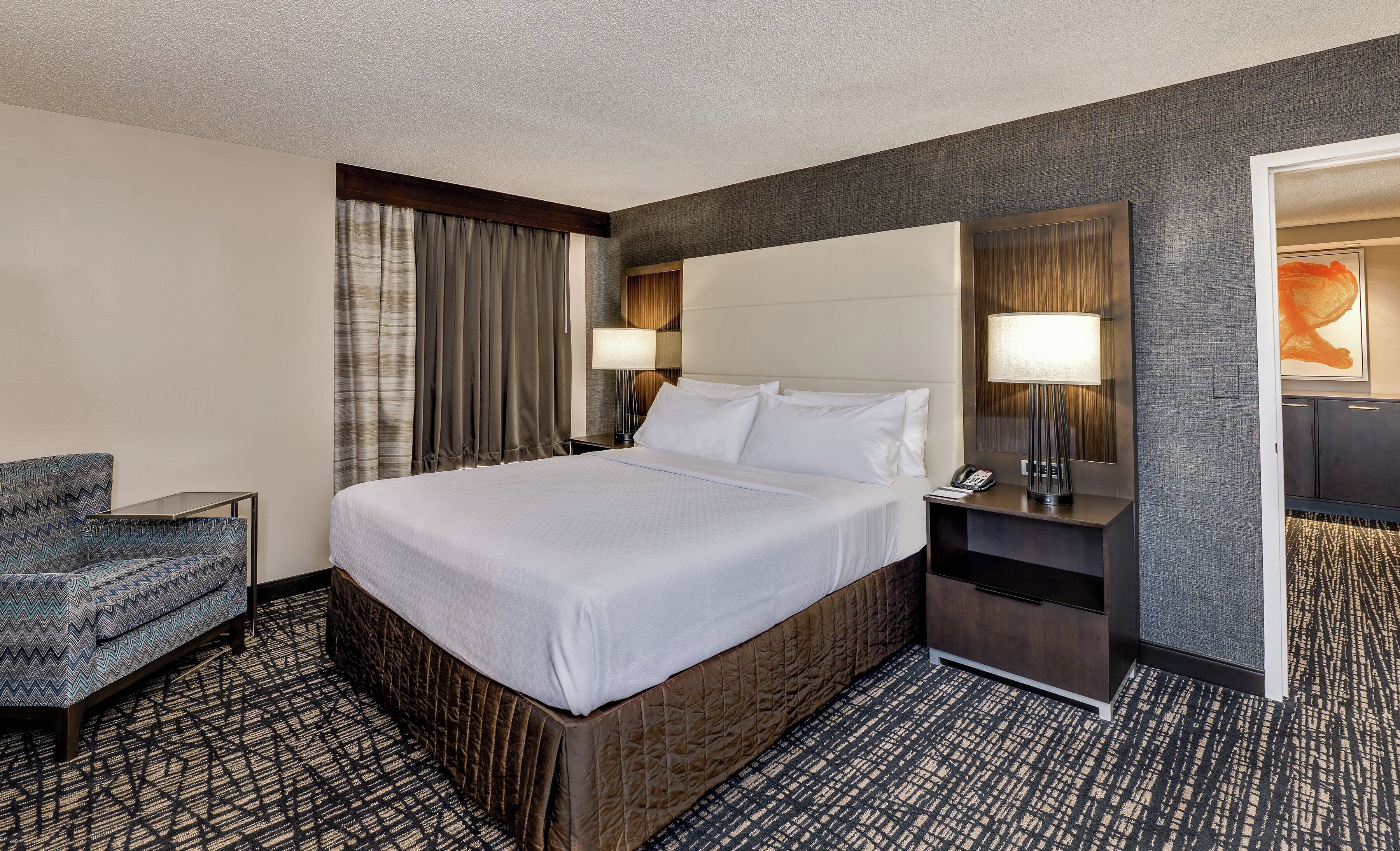 A hotel room with a large bed, a chair, a desk with a lamp, and a telephone.