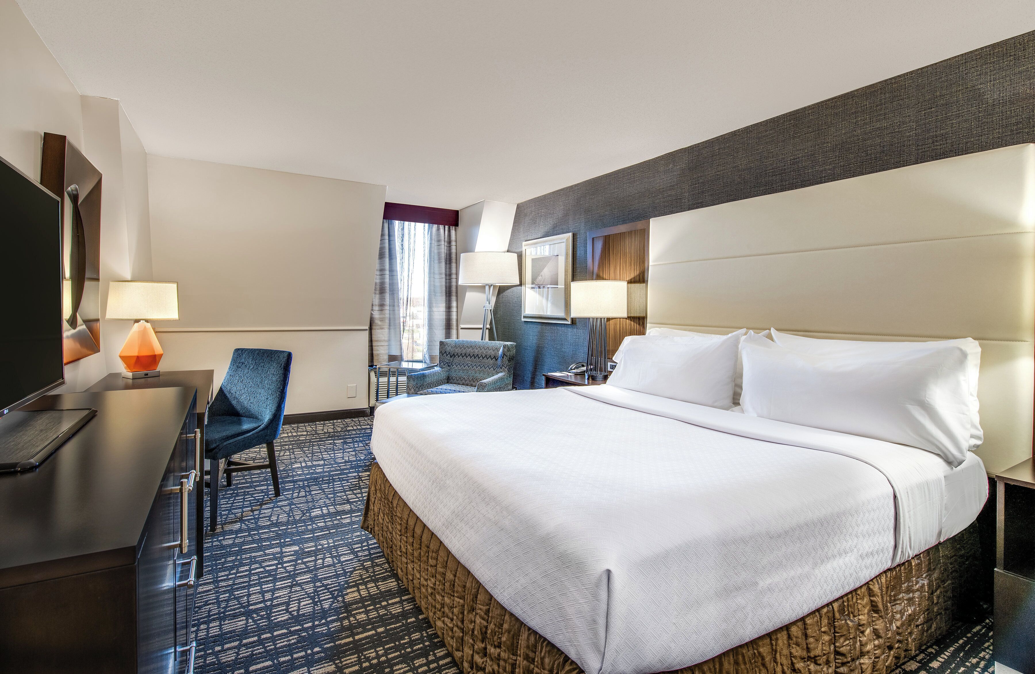 Ein Hotelzimmer mit einem großen Bett, einem Schreibtisch mit Stuhl, einem Fernseher und einem Fenster mit Vorhängen.