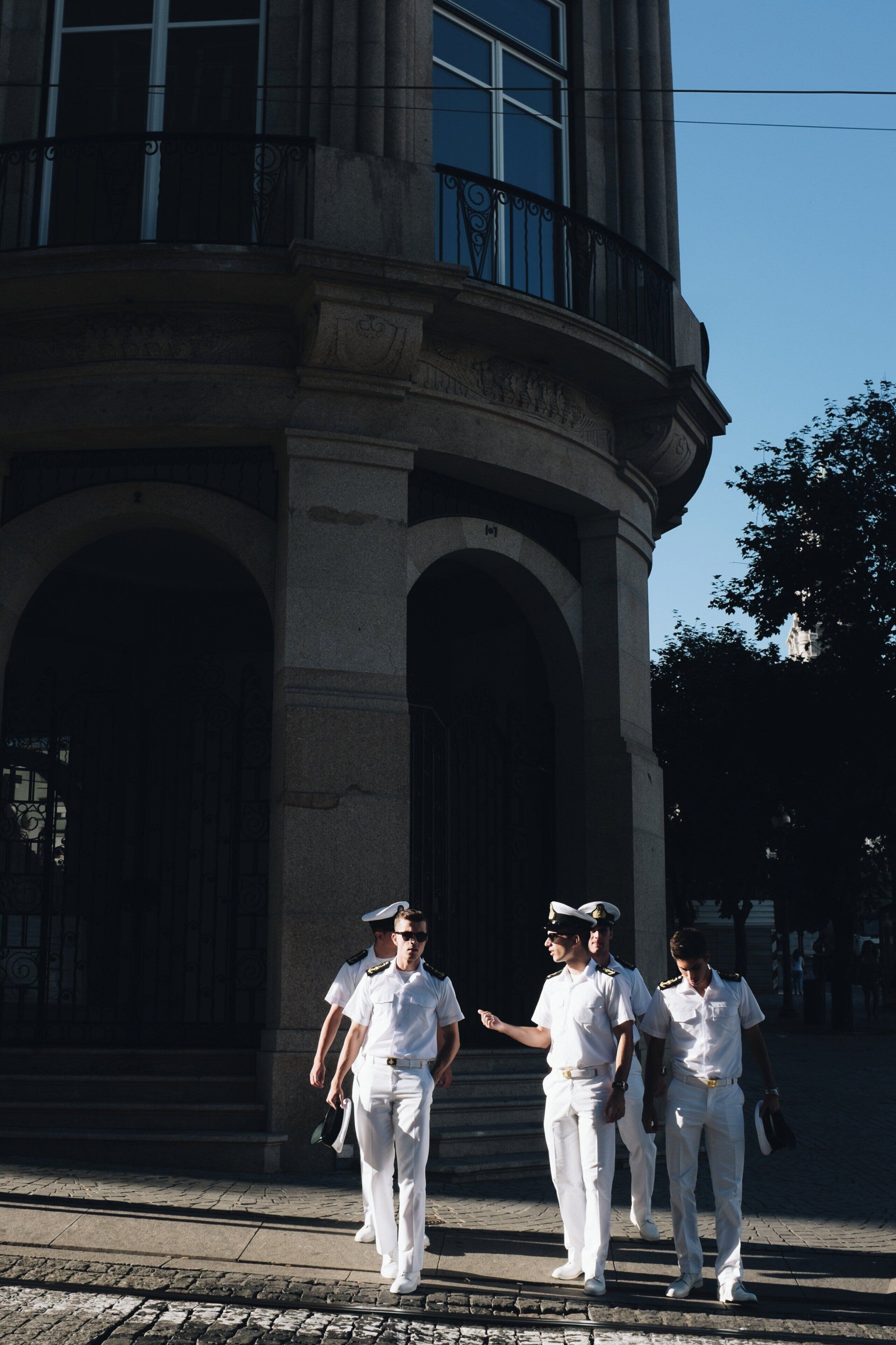 A group of uniformed individuals walking past a classical building with arched doorways and a balcony.