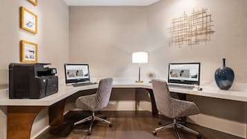 Um espaço de escritório moderno com duas mesas, cada uma com um computador e uma impressora. Há duas cadeiras, um vaso decorativo e quadros nas paredes.