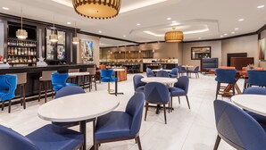 Um hall de hotel moderno com mesas redondas e cadeiras azuis, área de bar com diversas garrafas de bebidas e luminárias contemporâneas.