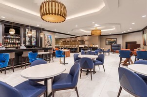 Un moderno vestíbulo de hotel con mesas redondas y sillas azules, una zona de bar con varias botellas de licores, e iluminación contemporánea.