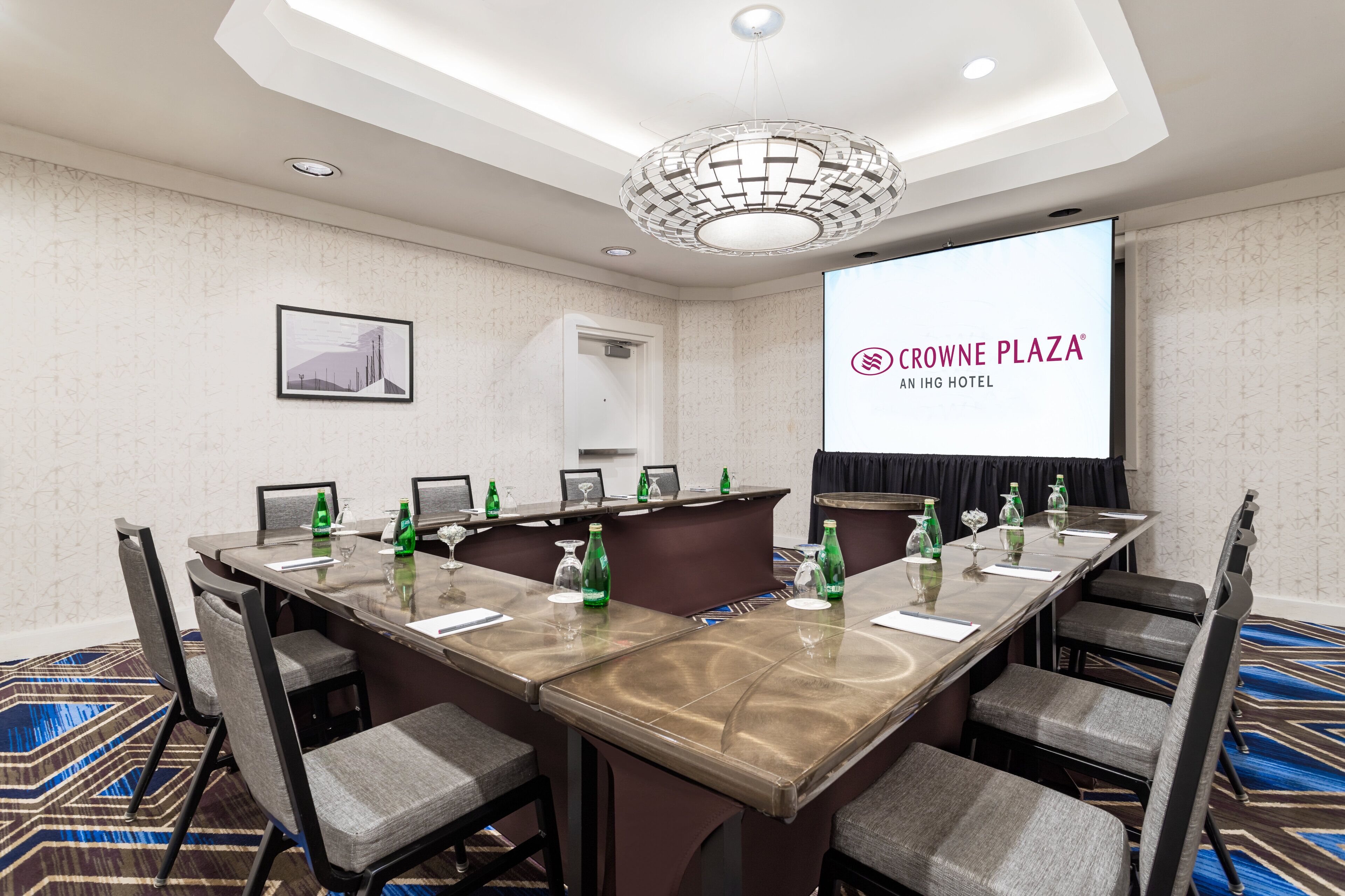 Un salón de conferencias con una larga mesa, sillas, una pantalla de proyector mostrando “Crowne Plaza An IHG Hotel” y una alfombra estampada.