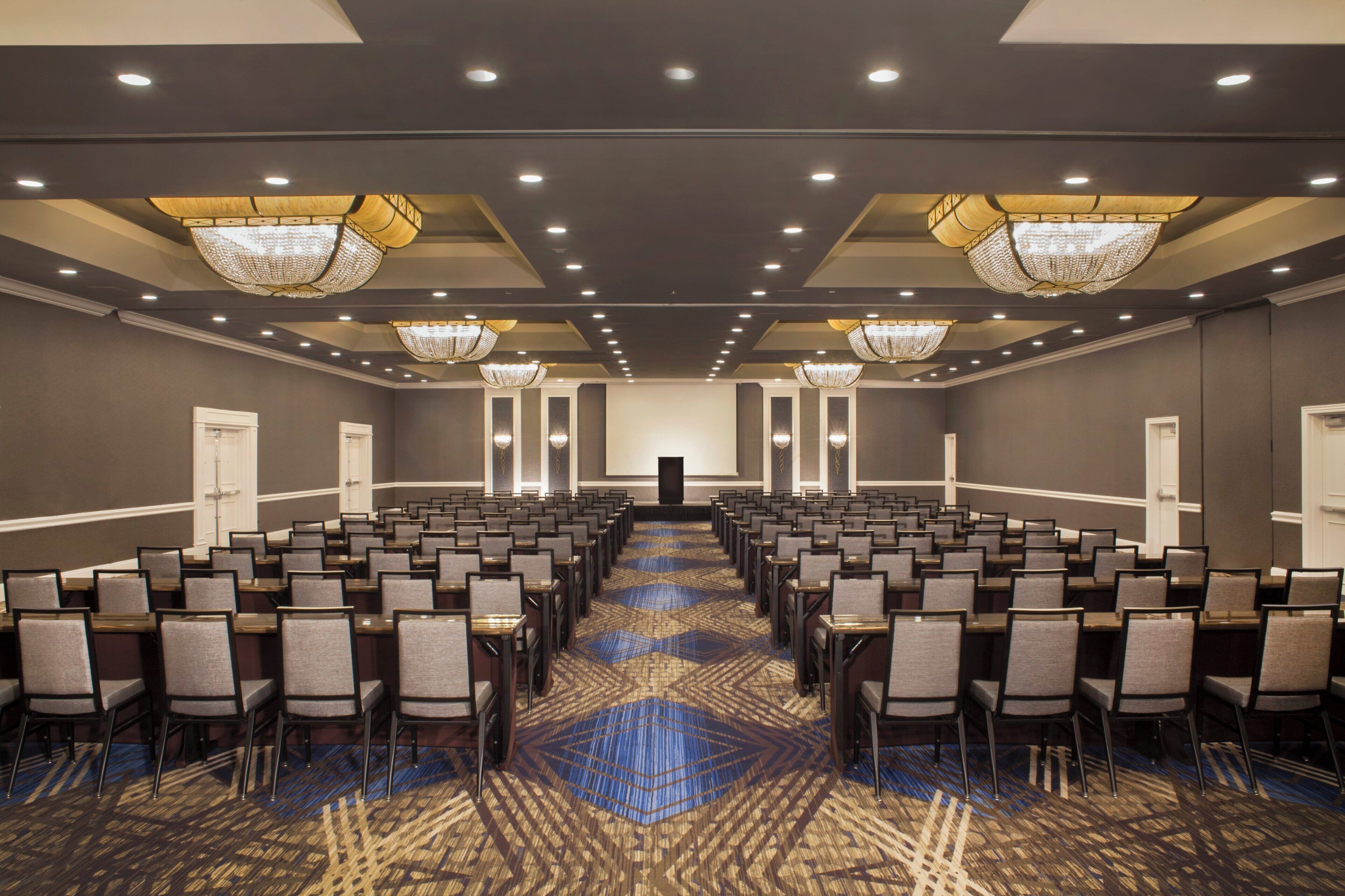 A conference room with rows of chairs, a patterned carpet, and chandeliers.
