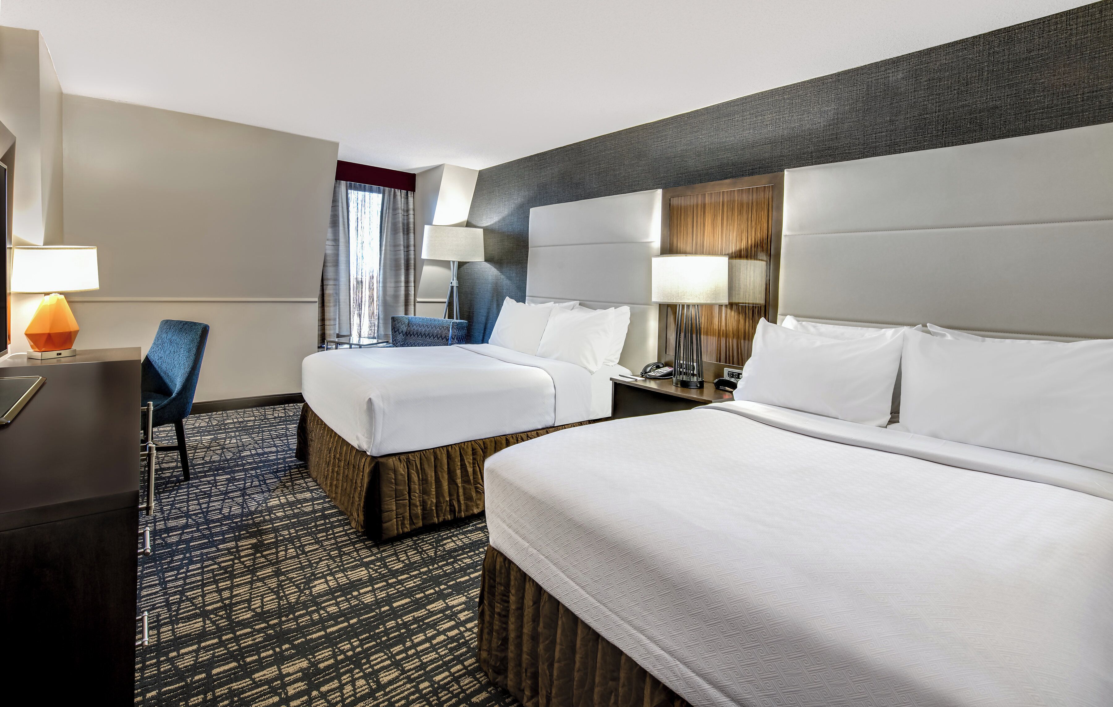 A hotel room with two beds, a desk, a chair, and a lamp.