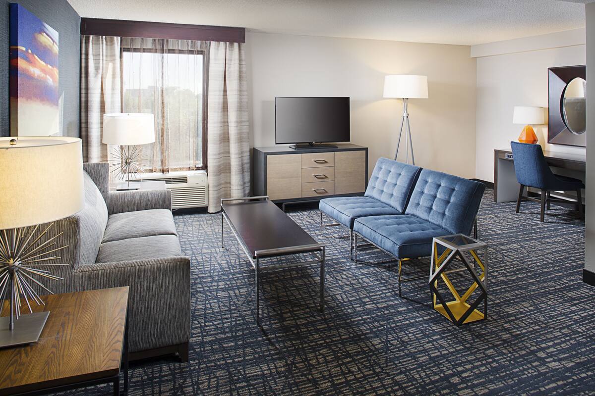 Ein Hotelzimmer mit einer Couch, einem Flachbildfernseher, einem Schreibtisch mit Stuhl, einer Lampe und einem gemusterten Teppich.