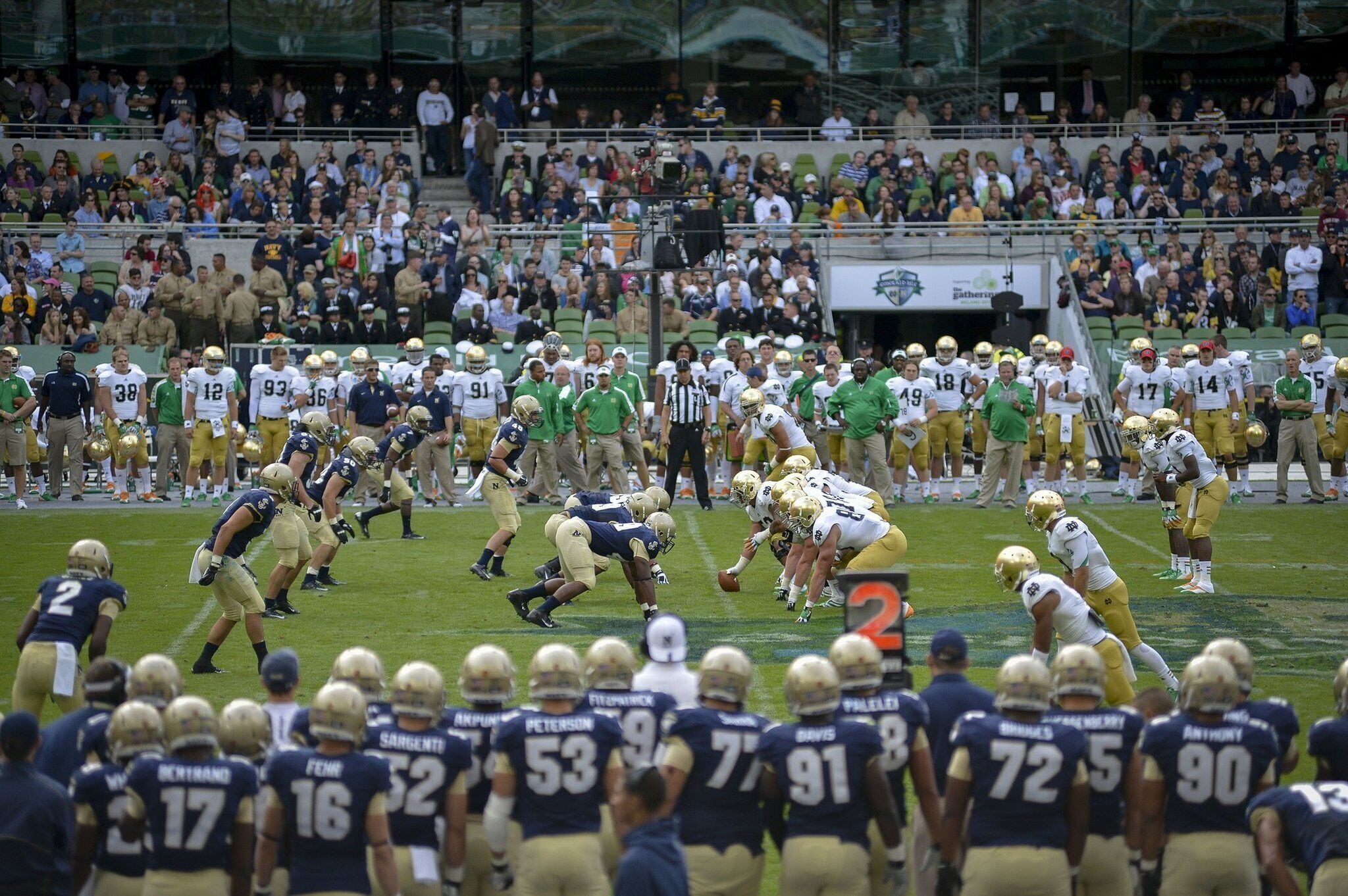 A football stadium with multiple teams on the field, spectators in the stands, and a scoreboard.
