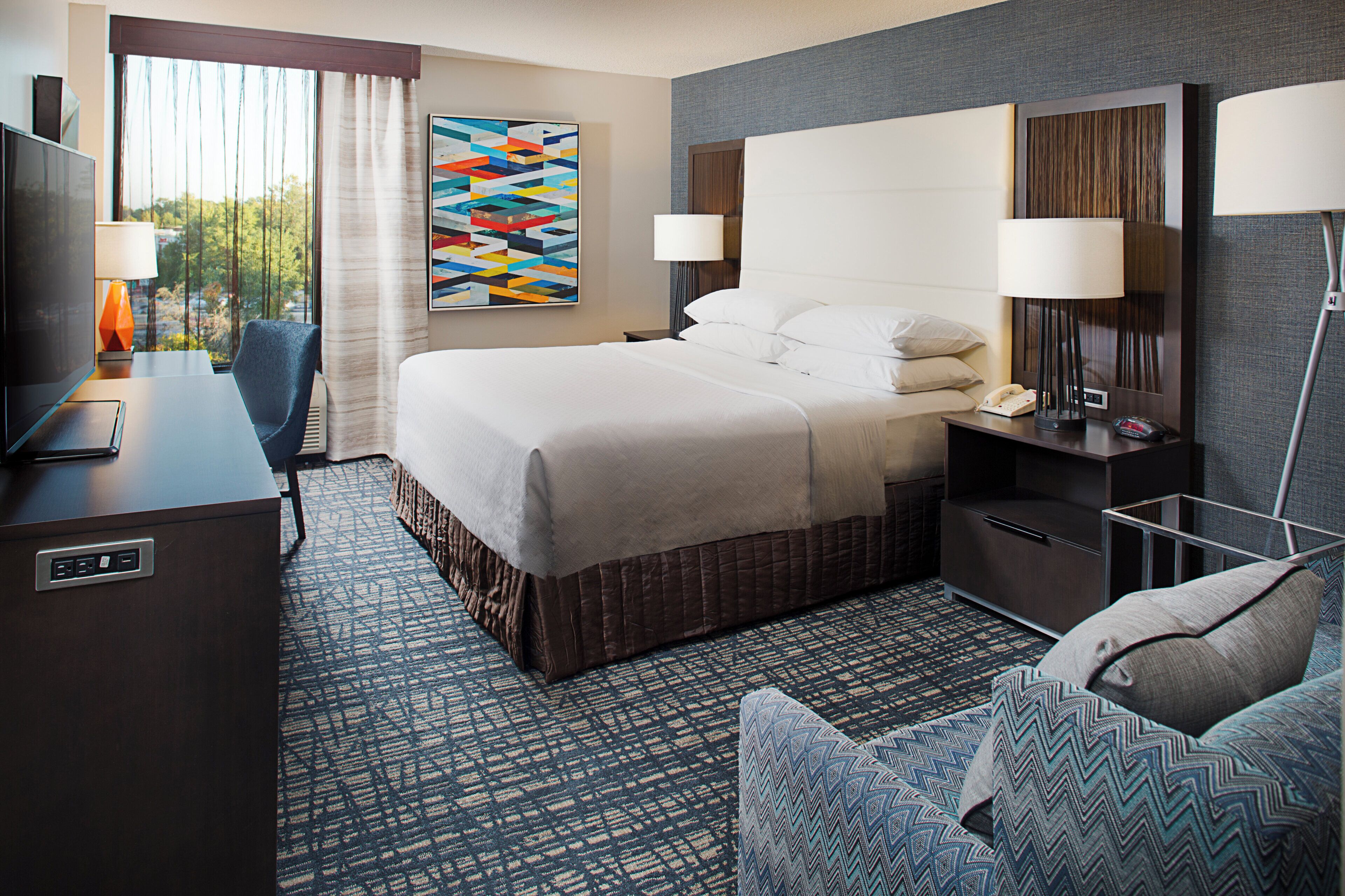 A hotel room with a bed, a desk with a TV, a chair, a bedside table with a lamp, and a wall art piece.