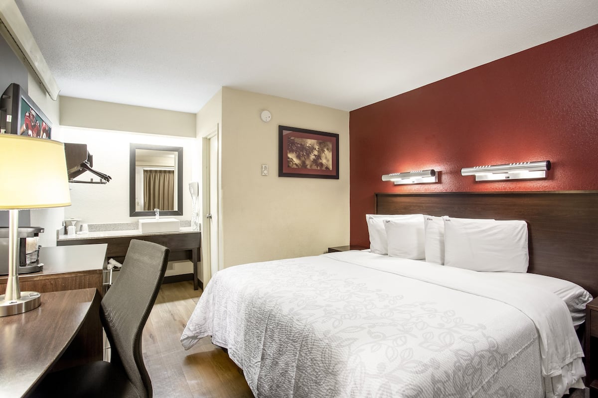 A hotel room with a bed, a desk with a lamp, a chair, a desk lamp, a mirror, a painting, and a wall-mounted light fixture.