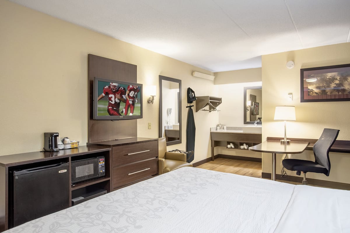 A hotel room with a bed, a TV, a desk with a chair, a microwave, a mini-fridge, and a bathroom with a sink and mirror.