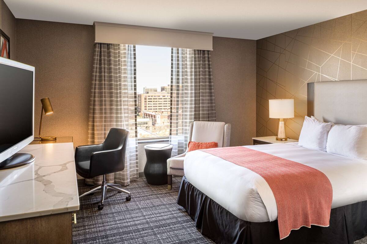 Ein Hotelzimmer mit einem großen Bett, zwei Nachttischen mit Lampen, einem Schreibtisch mit einem Computer, einem Sessel und Blick auf die Stadt durch große Fenster.