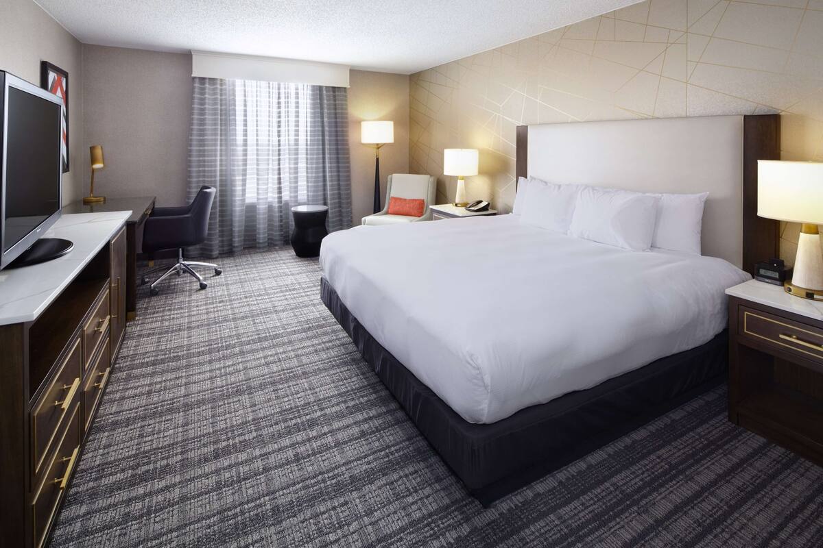 Ein Hotelzimmer mit einem großen Bett, einem Schreibtisch mit Computer, einem Sessel und einem Fernseher.