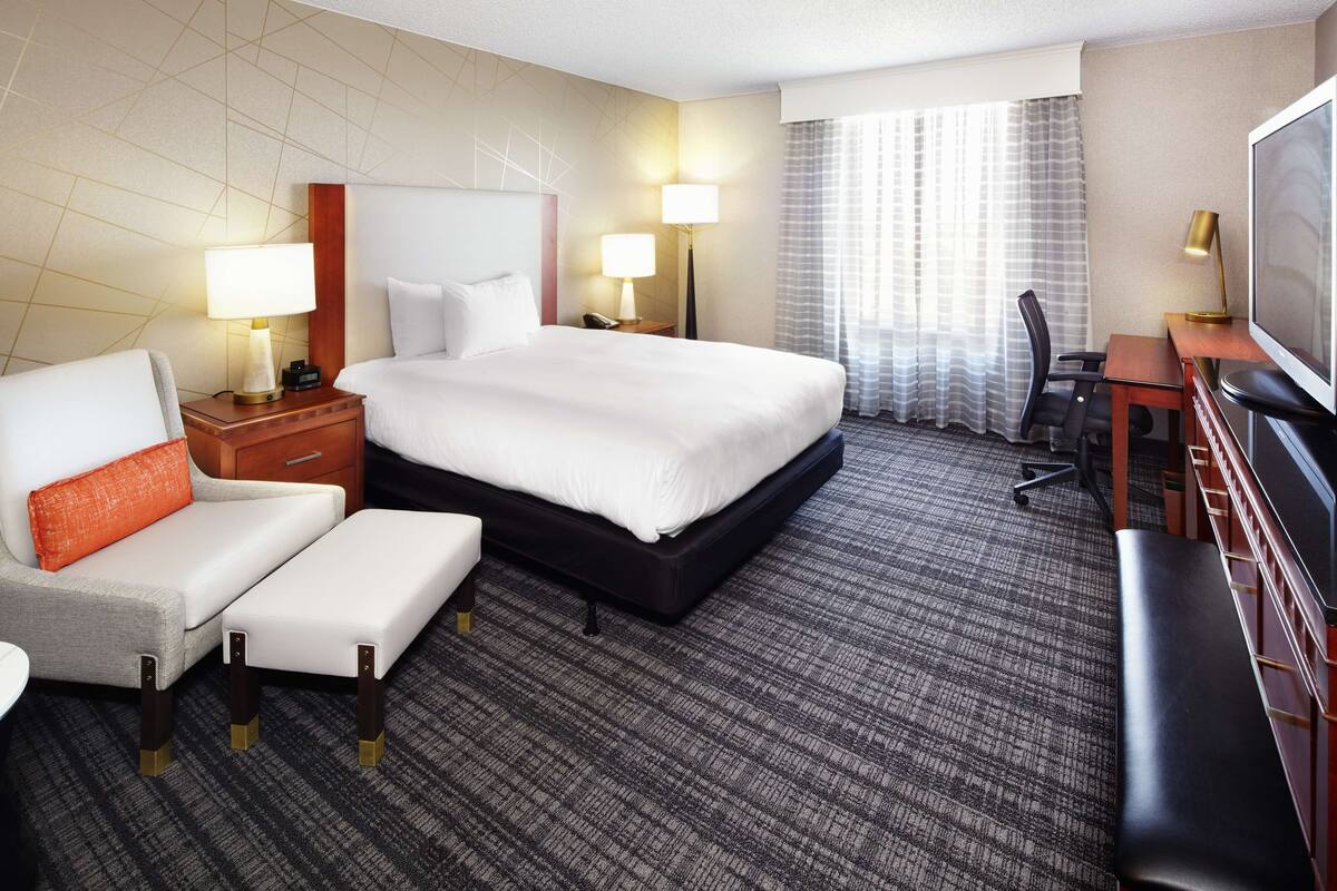 Ein Hotelzimmer mit einem großen Bett, einem Schreibtisch, einem Stuhl, einem Fernseher und einem Fenster mit Vorhängen.