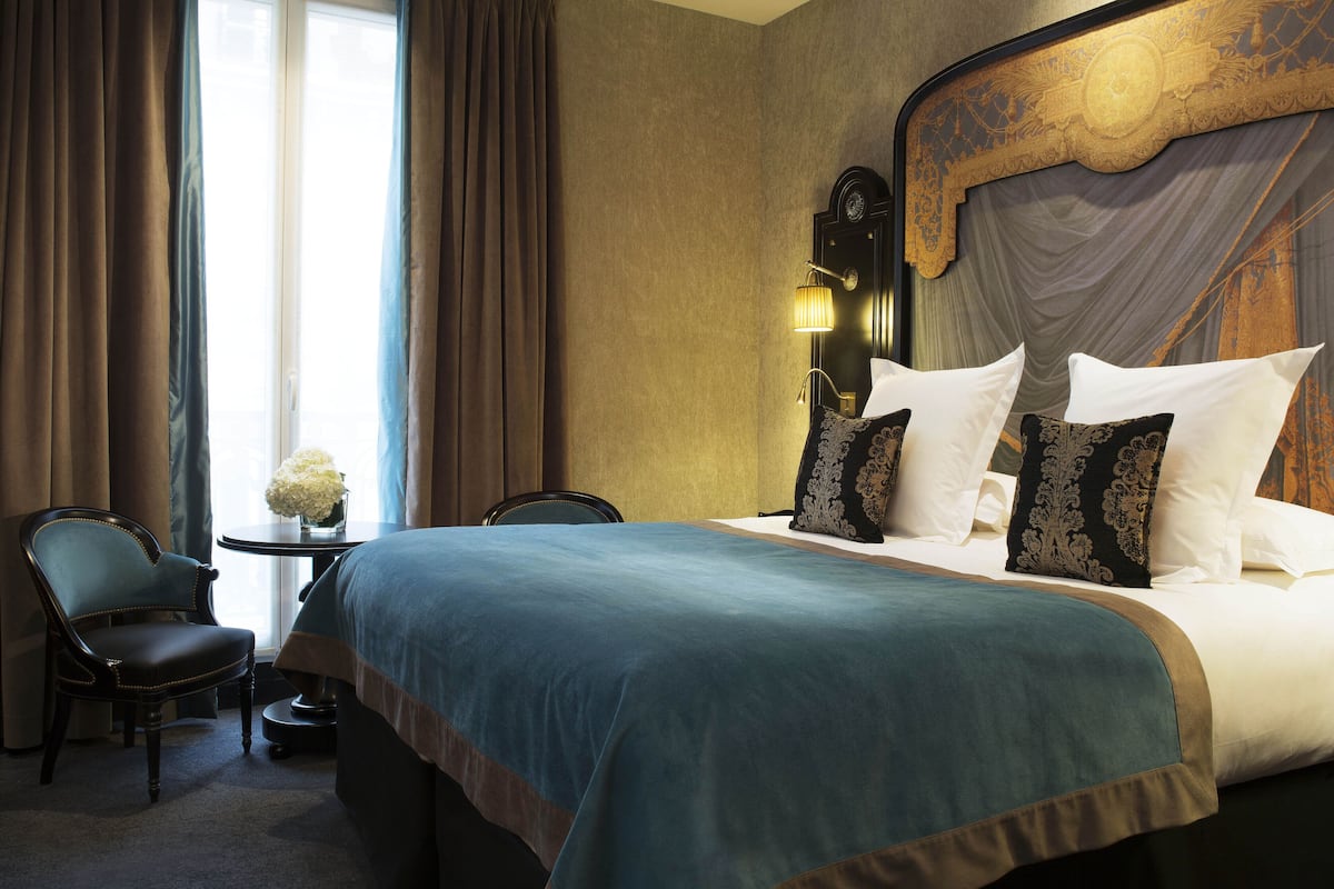 A hotel room with a large bed, two chairs, a small table with a vase, and a view of the outdoors.